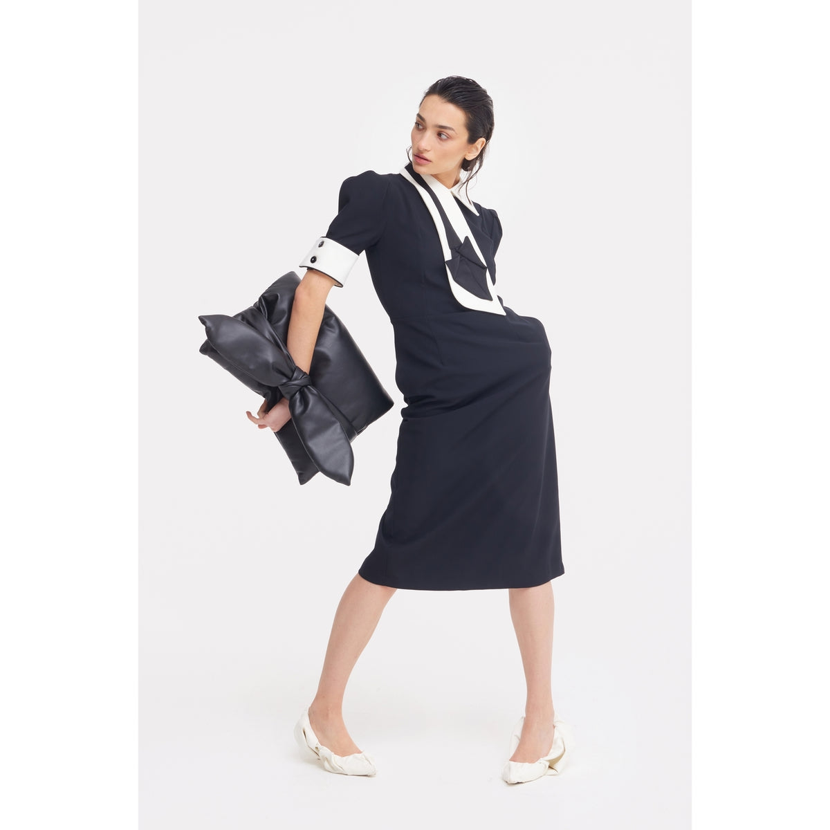 A woman in a black midi dress with white accents poses stylishly, holding the JULIA ALLERT Petal Faux Leather Evening Clutch Bag - Black. She wears white heels and has slicked-back hair, standing against a plain white background.