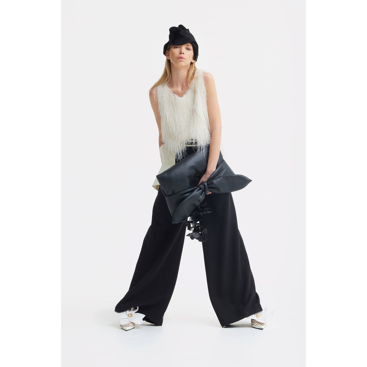 A woman poses in a sleeveless white fringe top, wide-leg black pants, embellished white pointed shoes, and a black hat by JULIA ALLERT. She holds an oversized black bow and carries the Petal Faux Leather Evening Clutch Bag in Black. The background is plain white.
