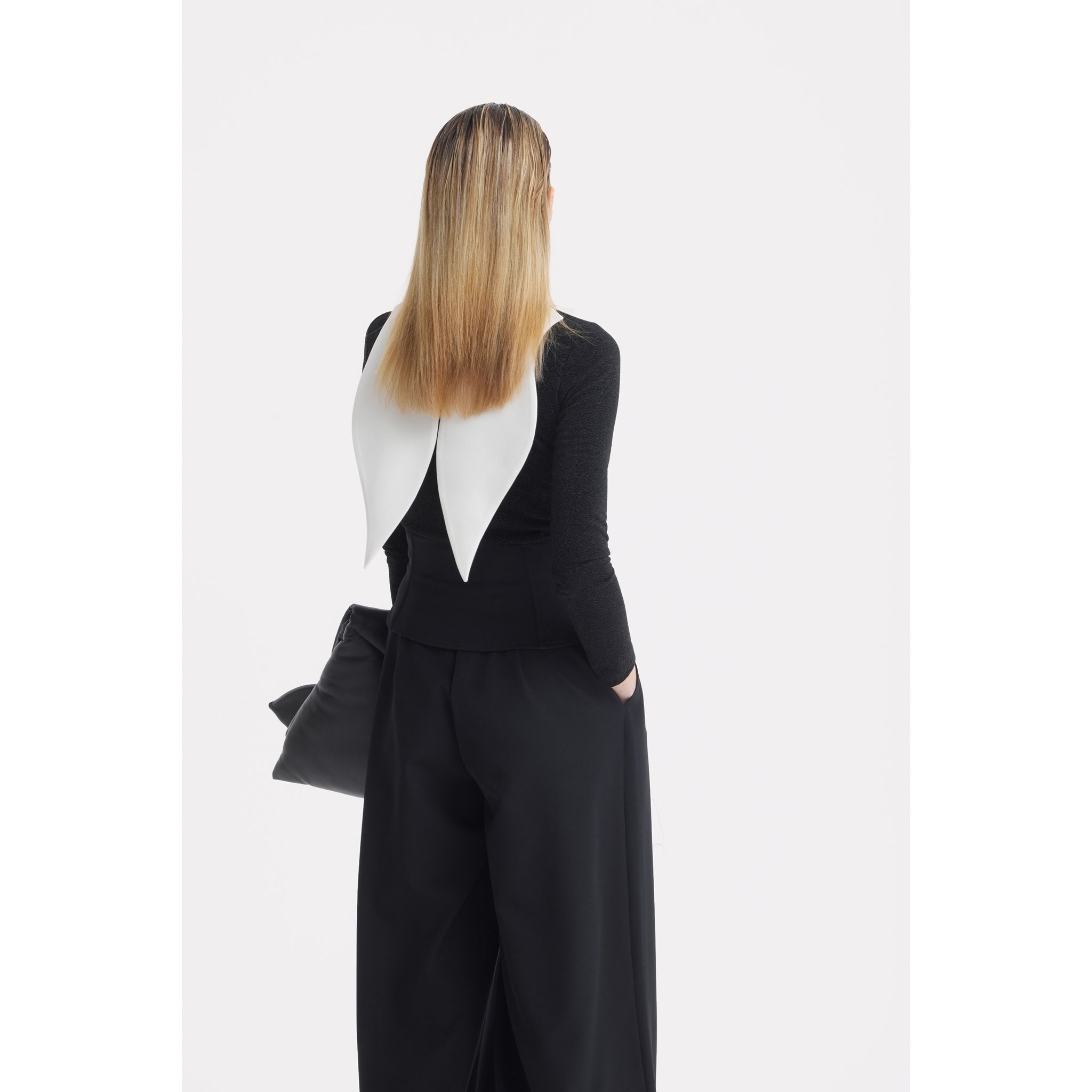 A person with straight, shoulder-length blonde hair stands facing away, dressed in black wide-leg pants and a top featuring the JULIA ALLERT Oversized Satin Bib Collar in Ivory. They are holding a black bag.