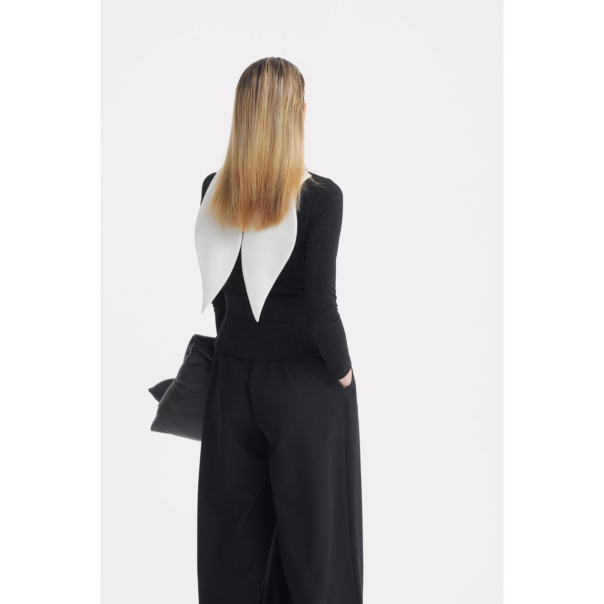 A person with straight, shoulder-length blonde hair stands facing away, dressed in black wide-leg pants and a top featuring the JULIA ALLERT Oversized Satin Bib Collar in Ivory. They are holding a black bag.