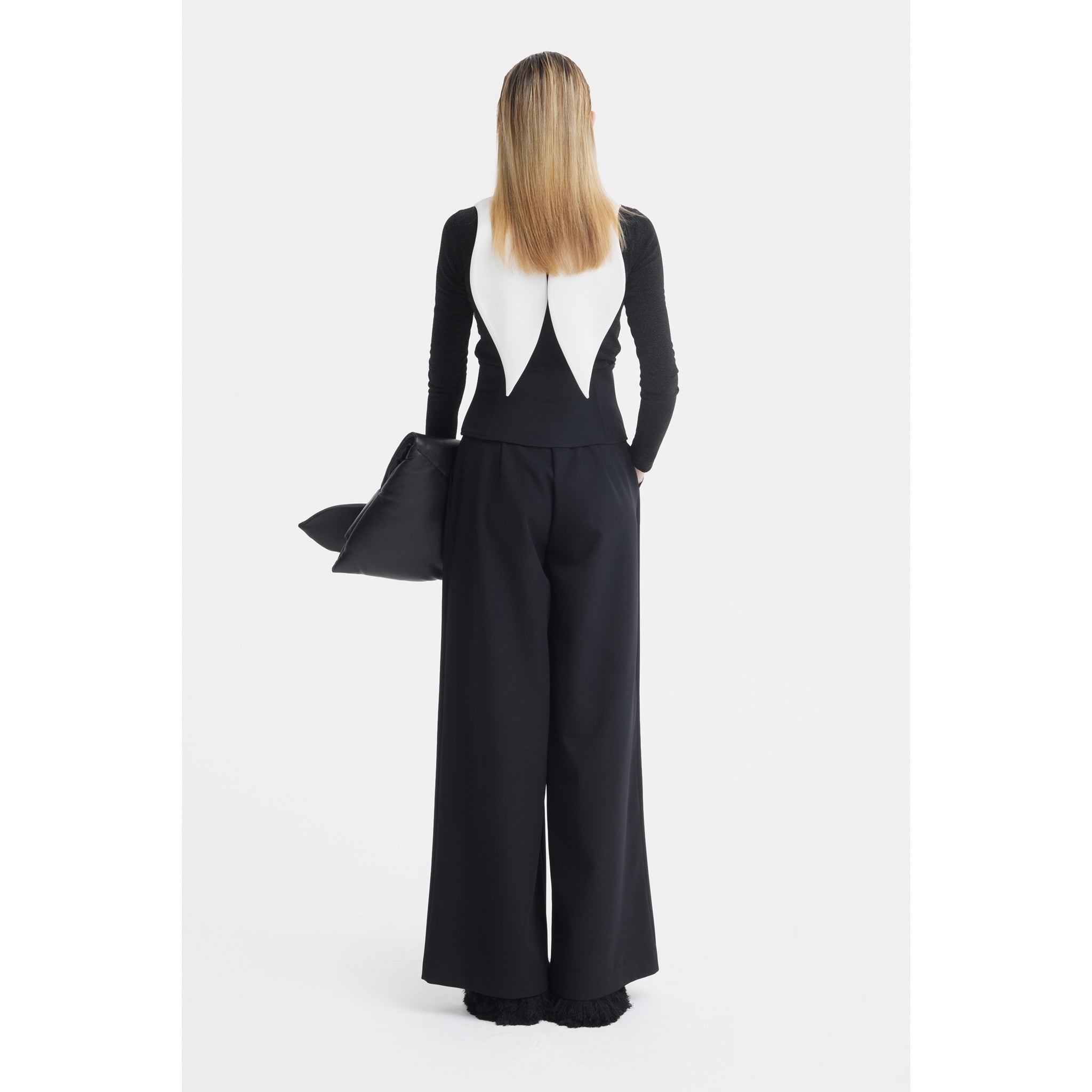 A person with straight blonde hair stands facing away, wearing JULIA ALLERT's Oversized Satin Bib Collar in ivory with a black long-sleeve top and wide-leg pants, holding a black clutch against a plain white background.