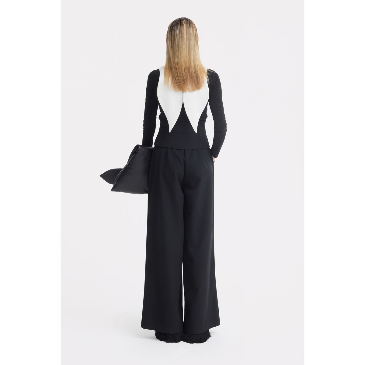 A person with straight blonde hair stands facing away, wearing JULIA ALLERT's Oversized Satin Bib Collar in ivory with a black long-sleeve top and wide-leg pants, holding a black clutch against a plain white background.