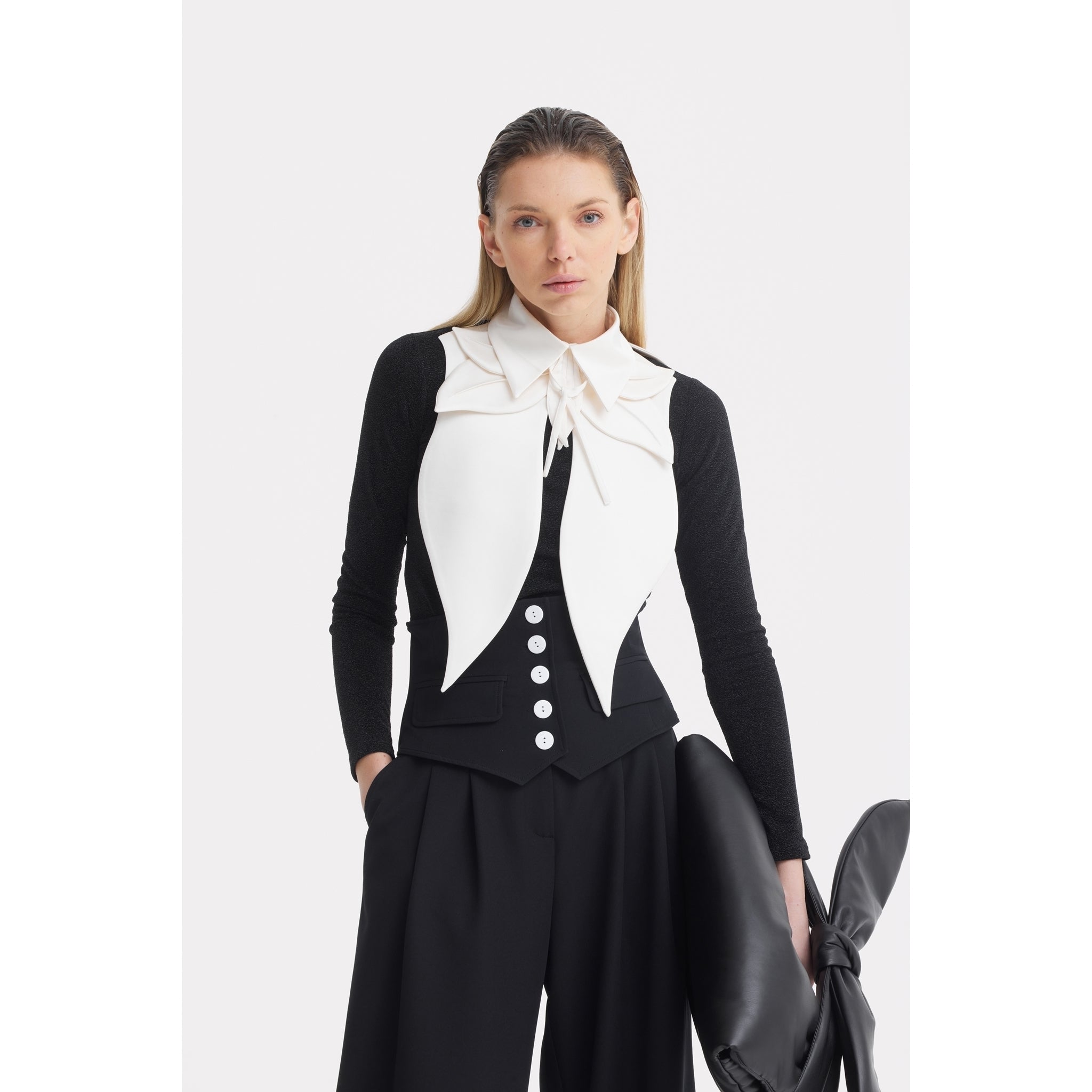 A woman with straight blonde hair poses in a black long-sleeve top and wide-leg pants, styled with the JULIA ALLERT Oversized Satin Bib Collar in Ivory. She also holds a large black accessory.