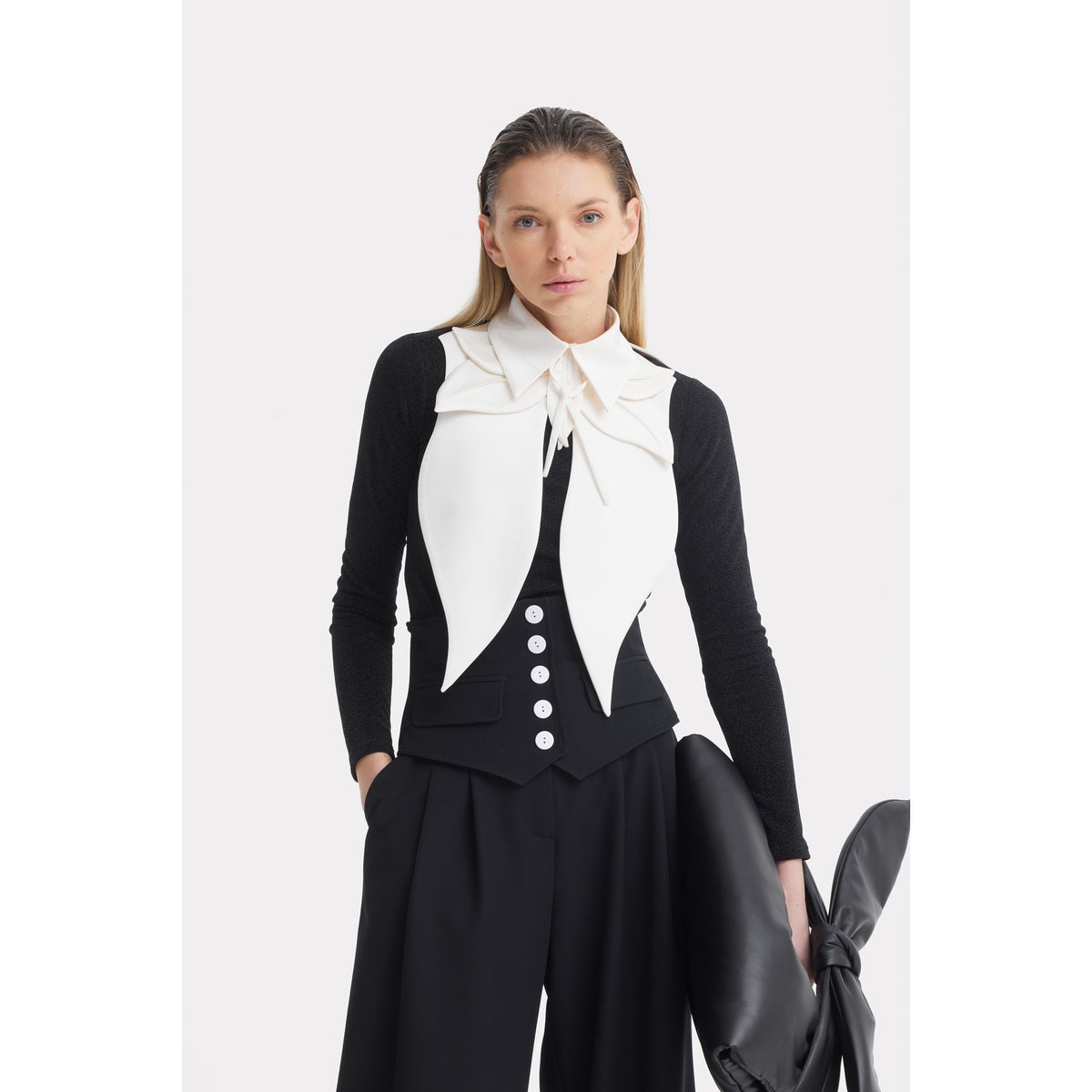 A woman with straight blonde hair poses in a black long-sleeve top and wide-leg pants, styled with the JULIA ALLERT Oversized Satin Bib Collar in Ivory. She also holds a large black accessory.