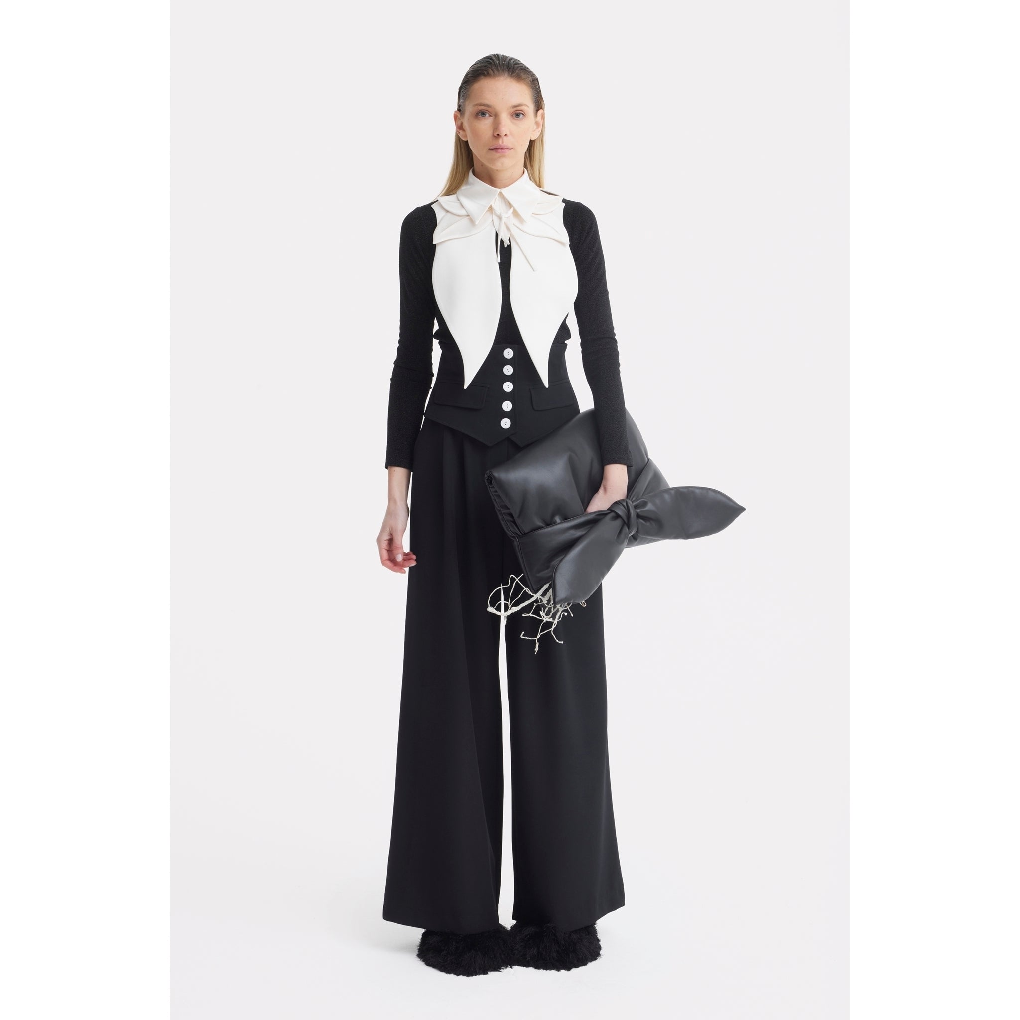 A woman stands against a white background wearing wide-leg black pants, a black top with the JULIA ALLERT Oversized Satin Bib Collar in ivory, and holds a large pillow-shaped clutch, her straight hair framing her neutral expression.