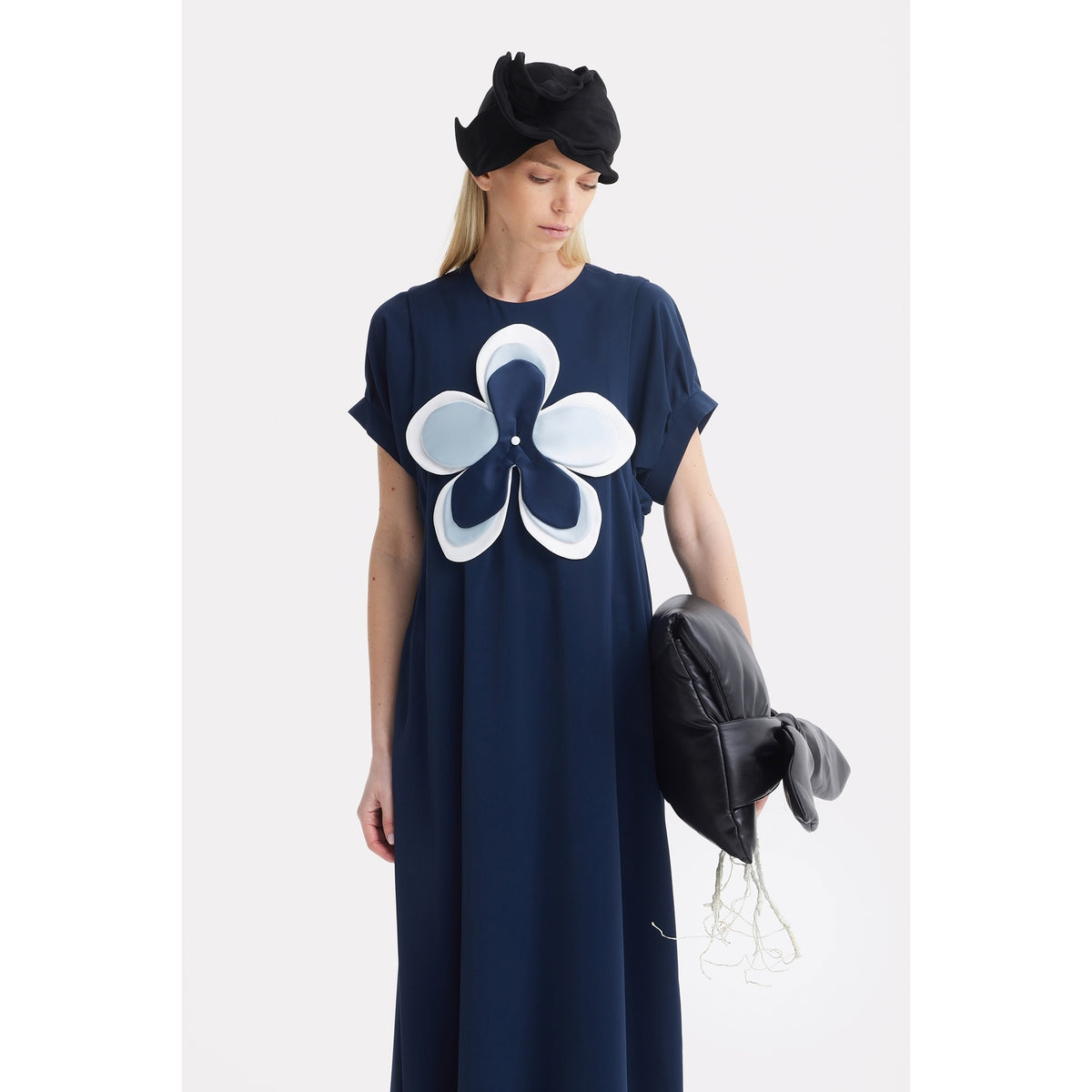 A woman in a navy blue dress with a bold white and blue floral design wears the JULIA ALLERT Headpiece - Evening Mini Hat Style in black, holds a black bow-accented bag, and stands against a plain white background, gazing down.