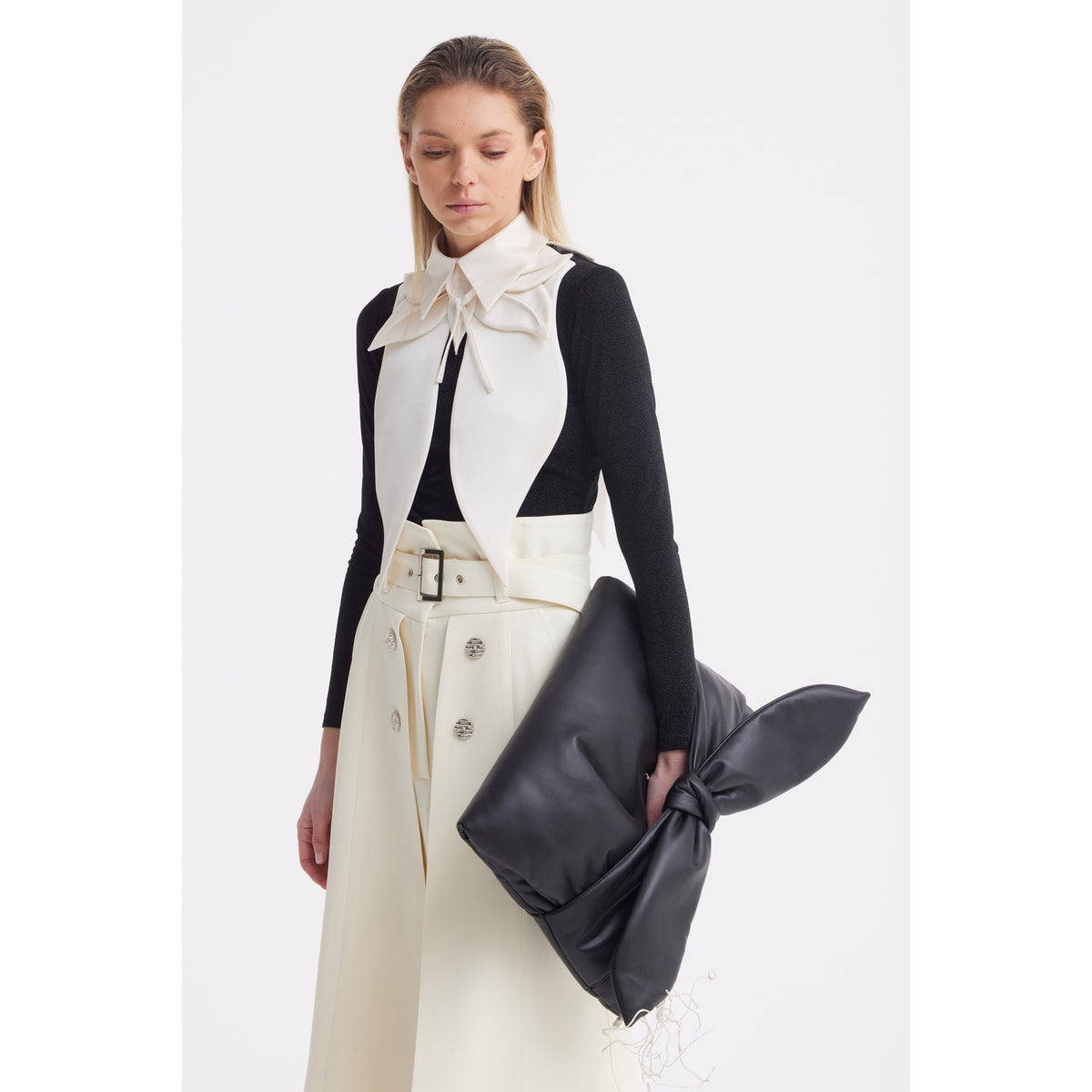 A woman in a black long-sleeve top, high-waisted white skirt, and the JULIA ALLERT Oversized Satin Bib Collar in ivory holds a large black clutch with a knot detail, standing against a plain white background.