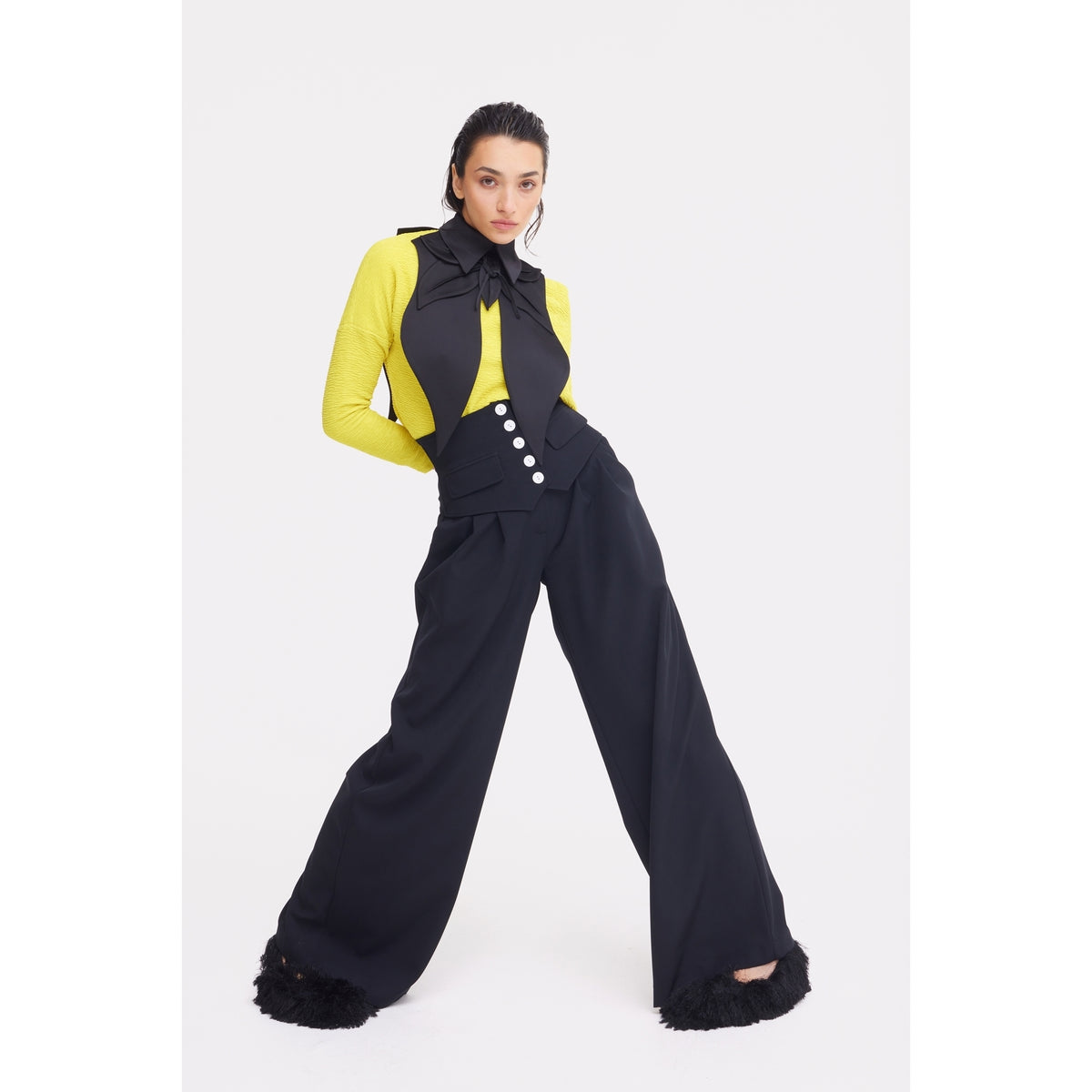 A person poses in JULIA ALLERT's Buttoned Corset-Waist Black Tailored Trousers, styled with a bright yellow top, a large black bow tie, and vest. The look features white buttons and sculpted details against a plain white background.