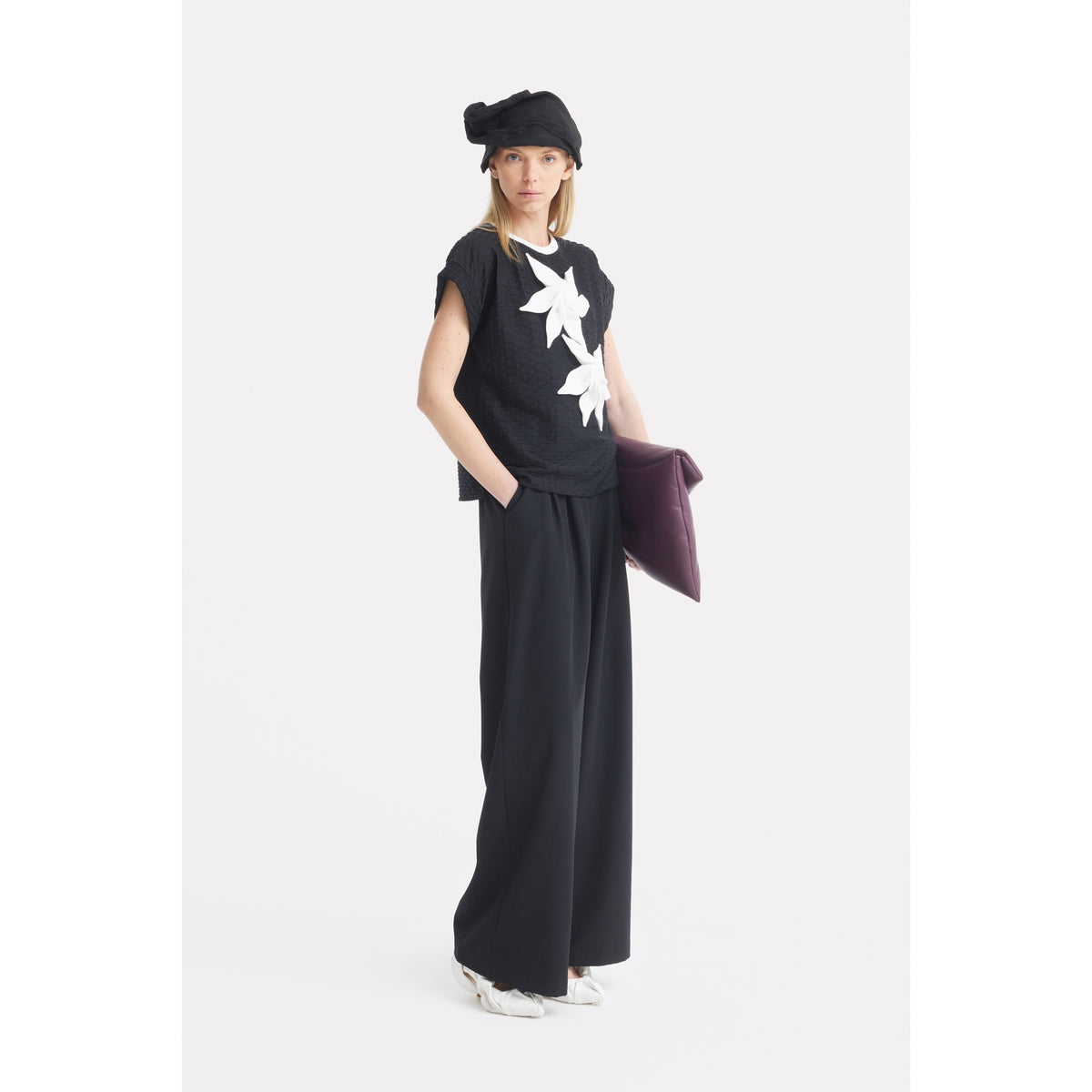 A woman poses against a white background, wearing a JULIA ALLERT Black Short Sleeve Top With Floral Detail, wide-leg black pants, black hat, and white shoes. She carries a large dark purple clutch under her arm with hands in her pockets.