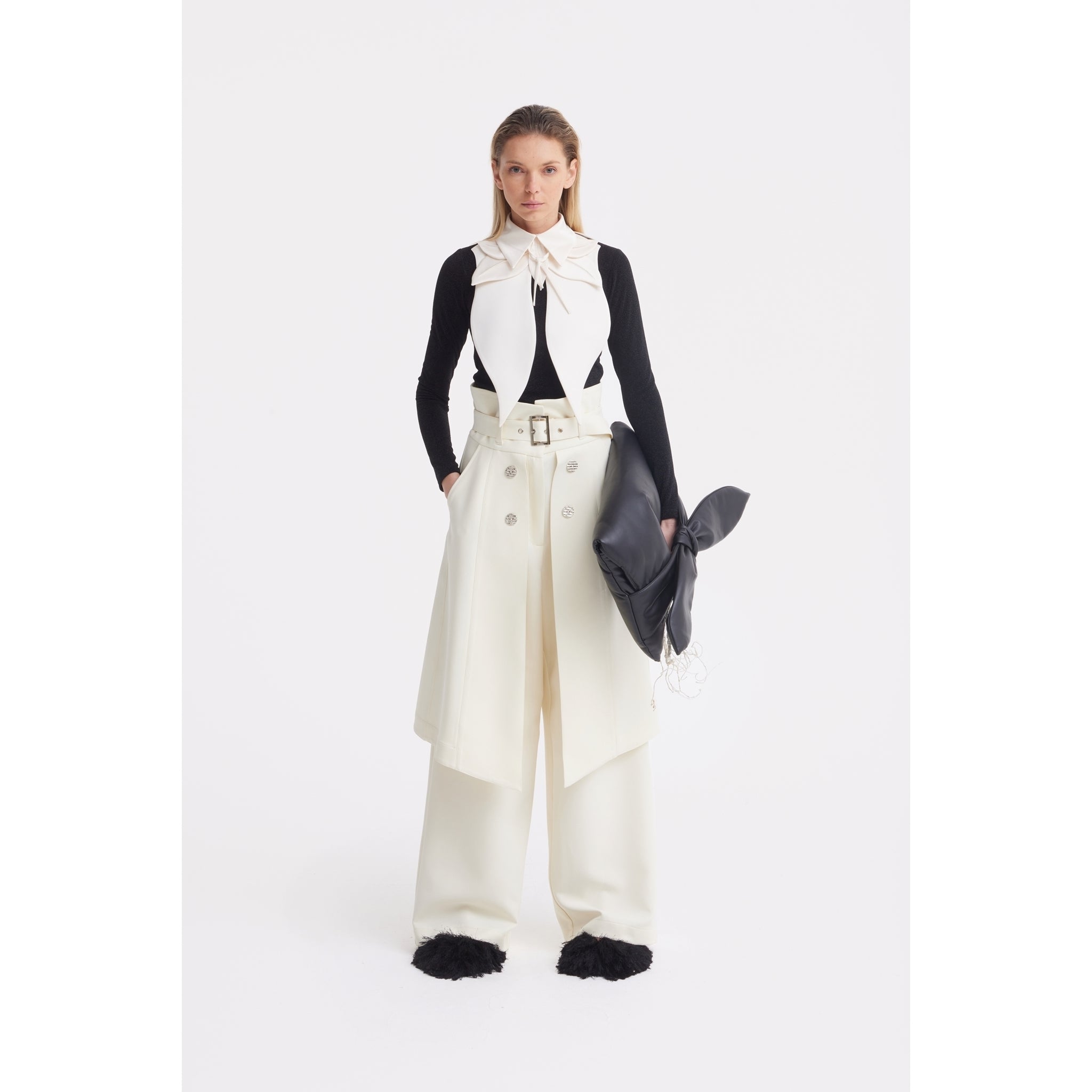 A woman with straight blonde hair stands against a white background, wearing cream wide-leg pants, a matching belted skirt, and the JULIA ALLERT Oversized Satin Bib Collar in ivory. She carries a large black clutch and wears a black long-sleeve top.