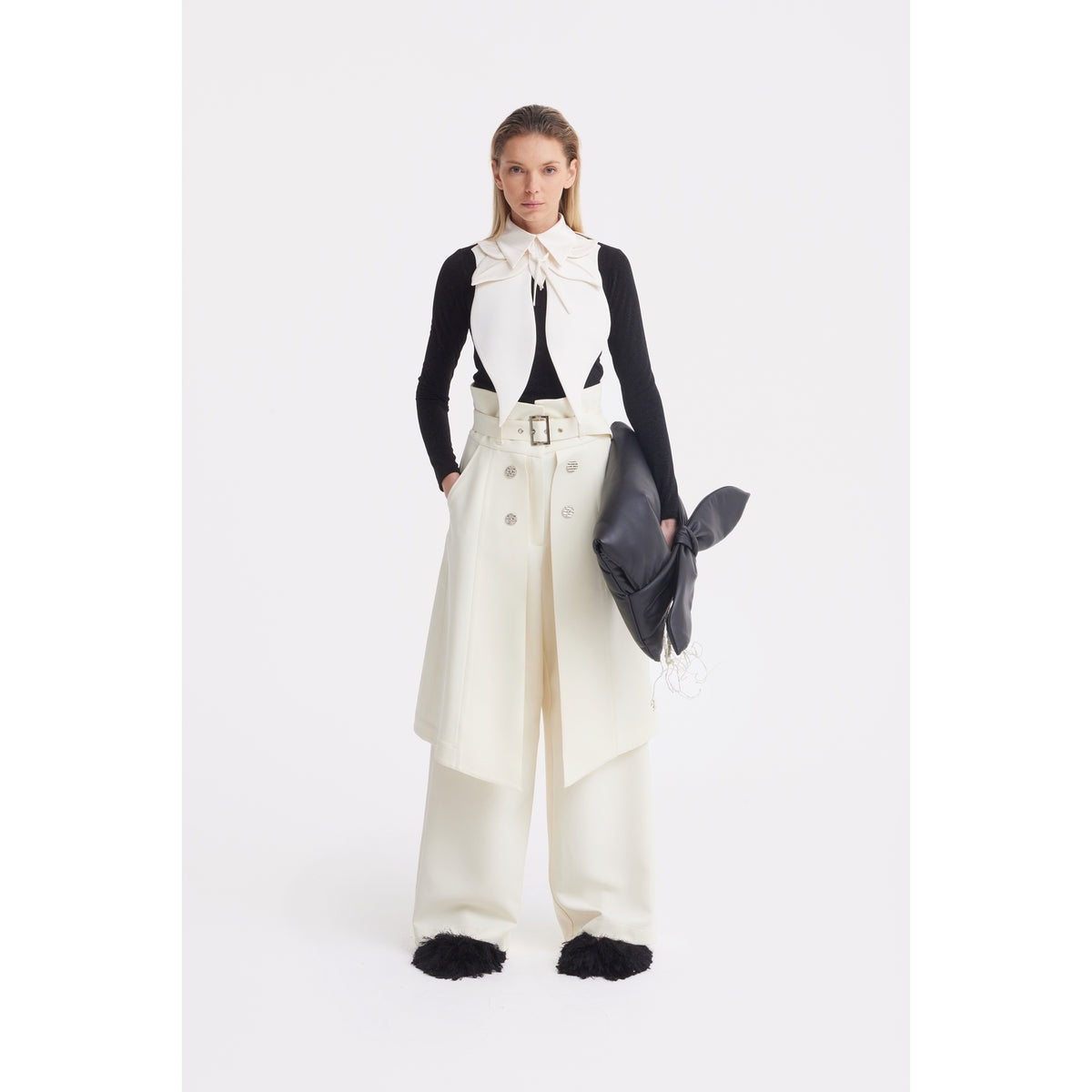 A woman with straight blonde hair stands against a white background, wearing cream wide-leg pants, a matching belted skirt, and the JULIA ALLERT Oversized Satin Bib Collar in ivory. She carries a large black clutch and wears a black long-sleeve top.