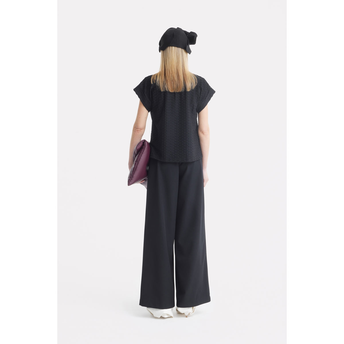 A person stands facing away against a white background, wearing a JULIA ALLERT Black Short Sleeve Top With Floral Detail, wide-leg black pants, a black bow hat, white shoes, and holding a large maroon clutch bag.