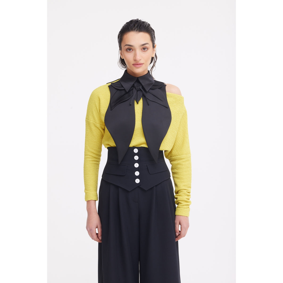 A woman stands facing forward in a yellow off-shoulder sweater, high-waisted black pants with white buttons, and the JULIA ALLERT Oversized Satin Bib Collar in black, her look elevated by this statement accessory against a plain white background.