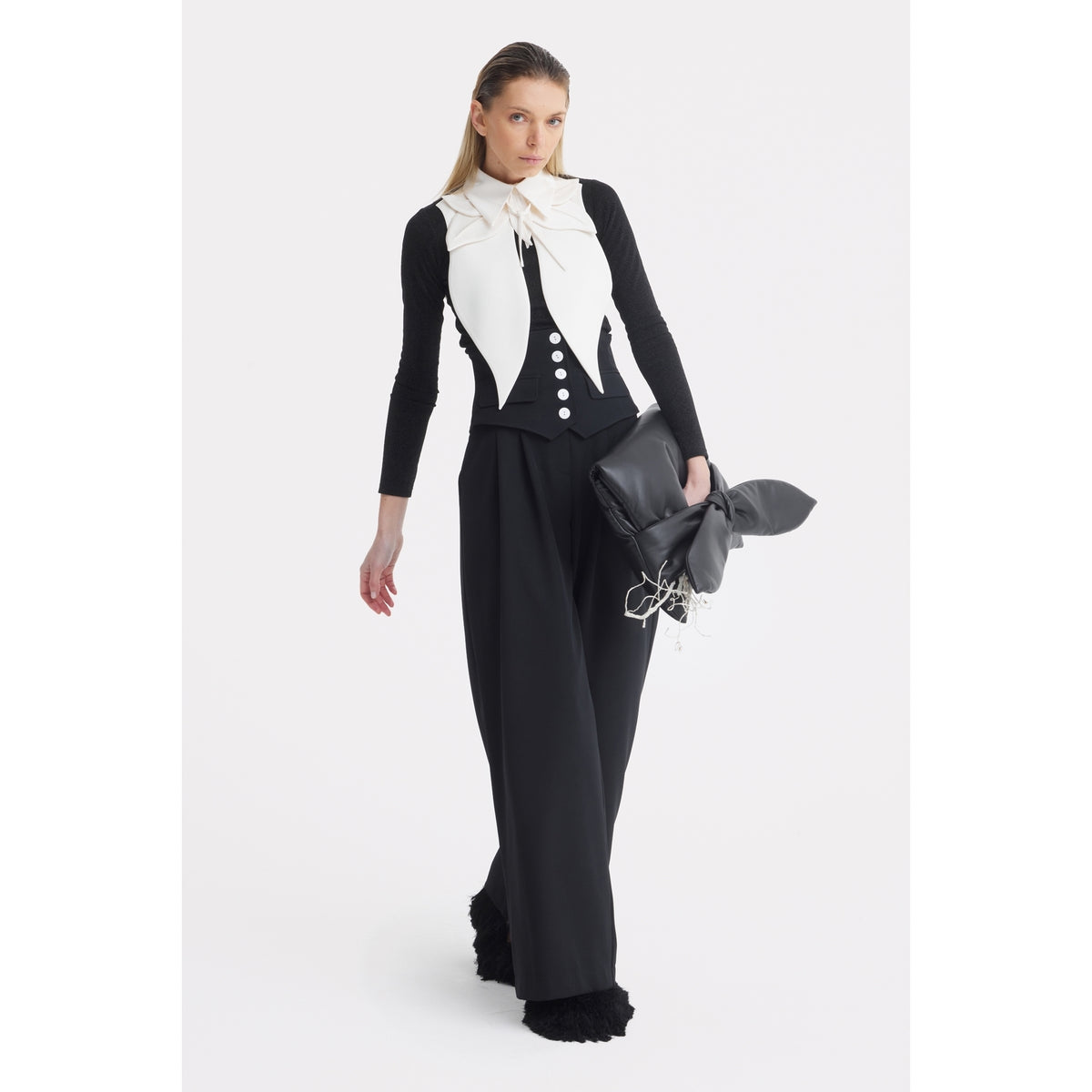 A woman models JULIA ALLERT’s Buttoned Corset-Waist Black Tailored Trousers with a black long-sleeve top and white vest-style overlay, carrying a large black bow clutch against a plain white background.