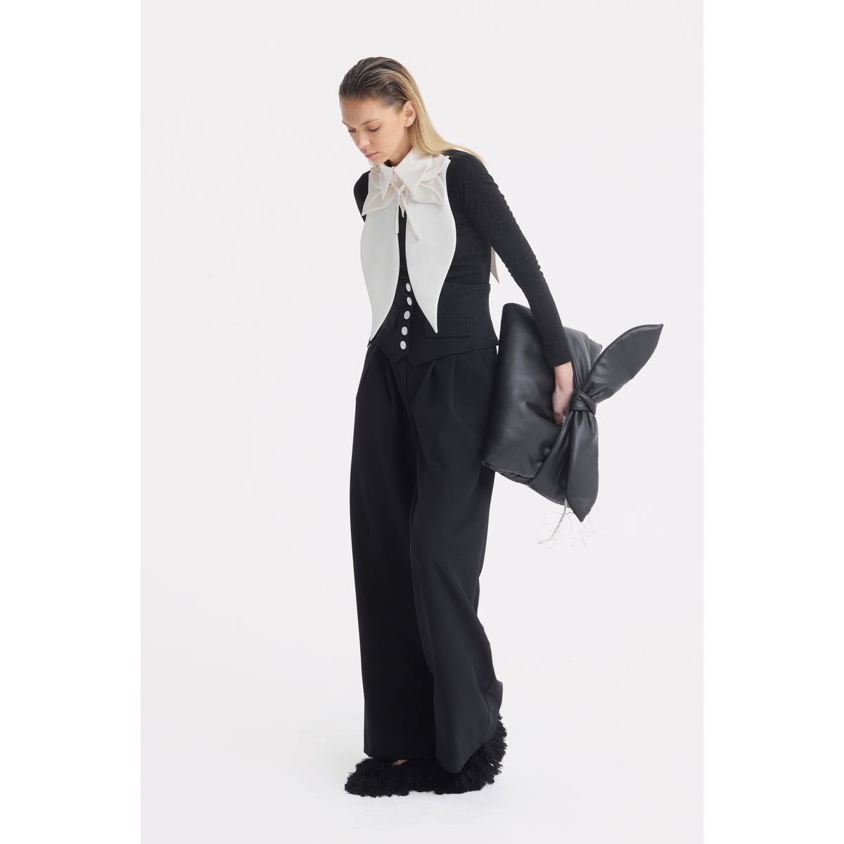 A woman in a black and white outfit holds a large black bunny pillow. She wears JULIA ALLERT's Buttoned Corset-Waist Black Tailored Trousers, a black top with white buttons, and a large white bow at her neck.