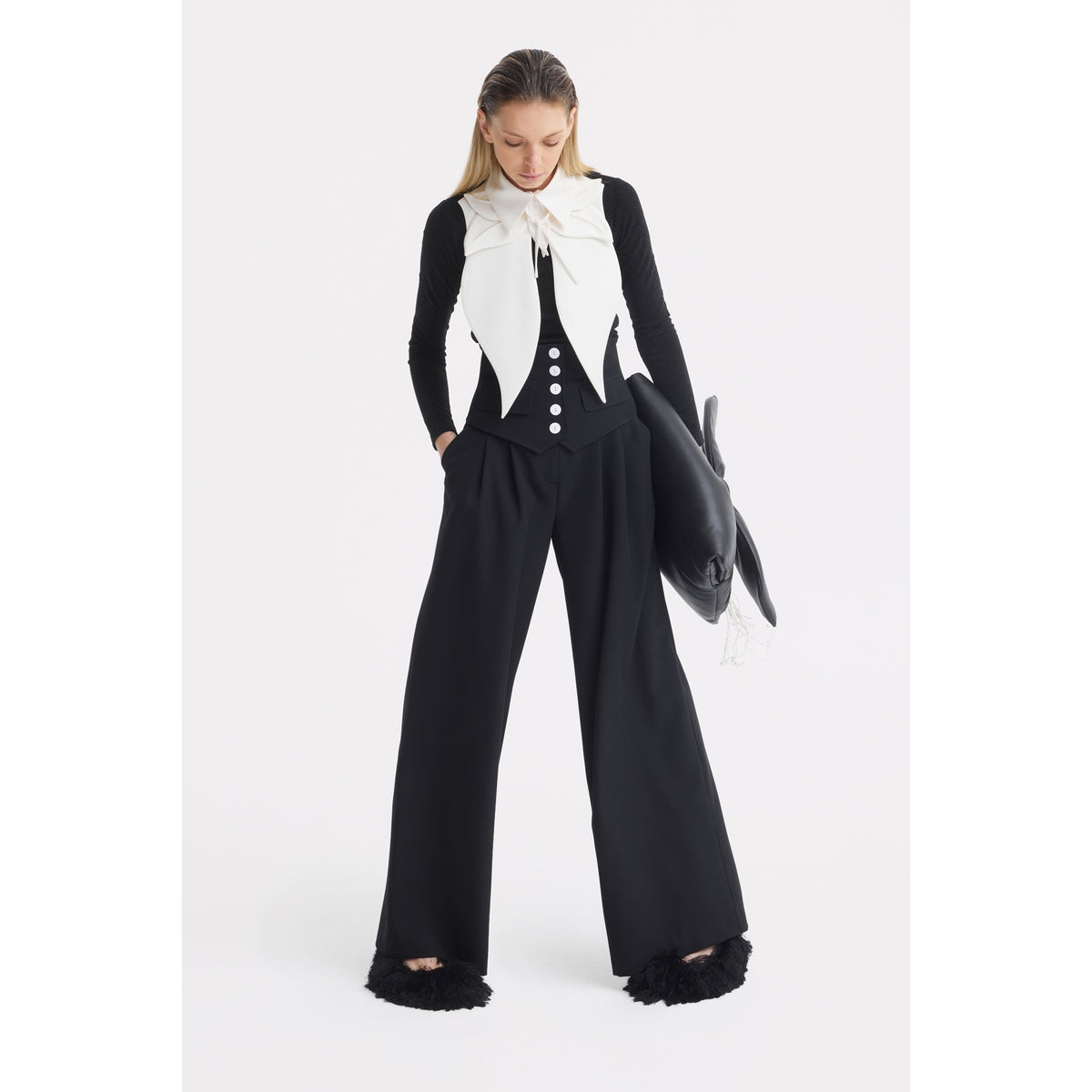 A woman stands against a white background, wearing JULIA ALLERT’s Buttoned Corset-Waist Black Tailored Trousers, paired with a fitted black top, a white sleeveless vest with large bow accents, and holding a black dolphin-shaped handbag.