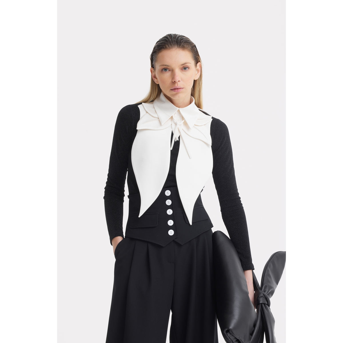 A woman with straight blonde hair wears JULIA ALLERT Buttoned Corset-Waist Black Tailored Trousers, a black long-sleeve top, a black vest with white buttons, and a white collar accessory, posing against a plain white background.