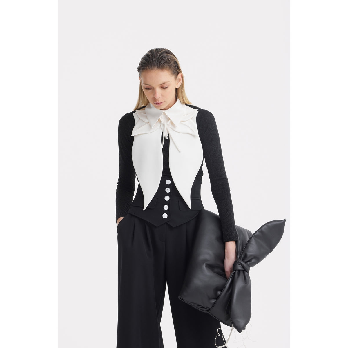 A woman with straight, light brown hair looks down, wearing a black long-sleeve top, white vest with a large bow collar, and JULIA ALLERT Buttoned Corset-Waist Black Tailored Trousers, holding a large black bow-shaped clutch.