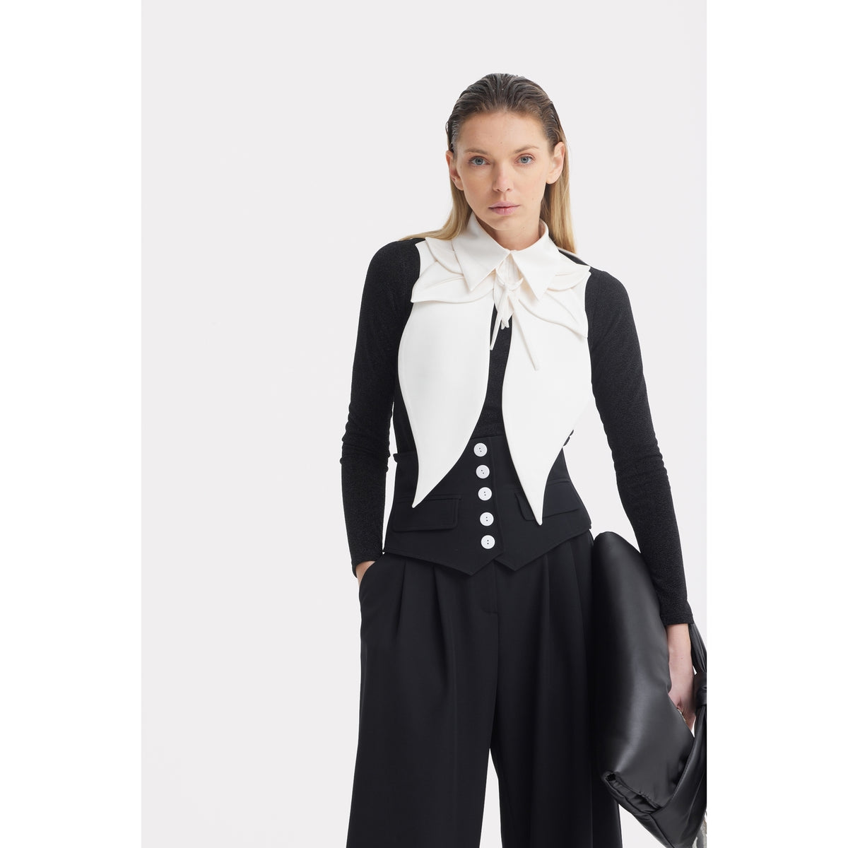A woman with straight blond hair wears a white sleeveless vest over a black long-sleeve top with a large white bow collar, JULIA ALLERT Buttoned Corset-Waist Black Tailored Trousers, and holds a large black clutch bag.