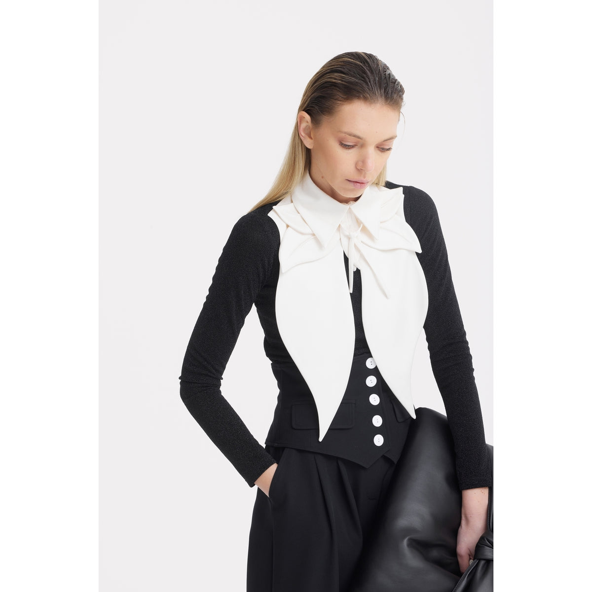 A woman with straight blonde hair wears JULIA ALLERT Buttoned Corset-Waist Black Tailored Trousers, a white bow collar, a fitted black vest with sculpted details and white buttons. She stands with one hand in her pocket, holding a large black bag, and looks down neutrally.