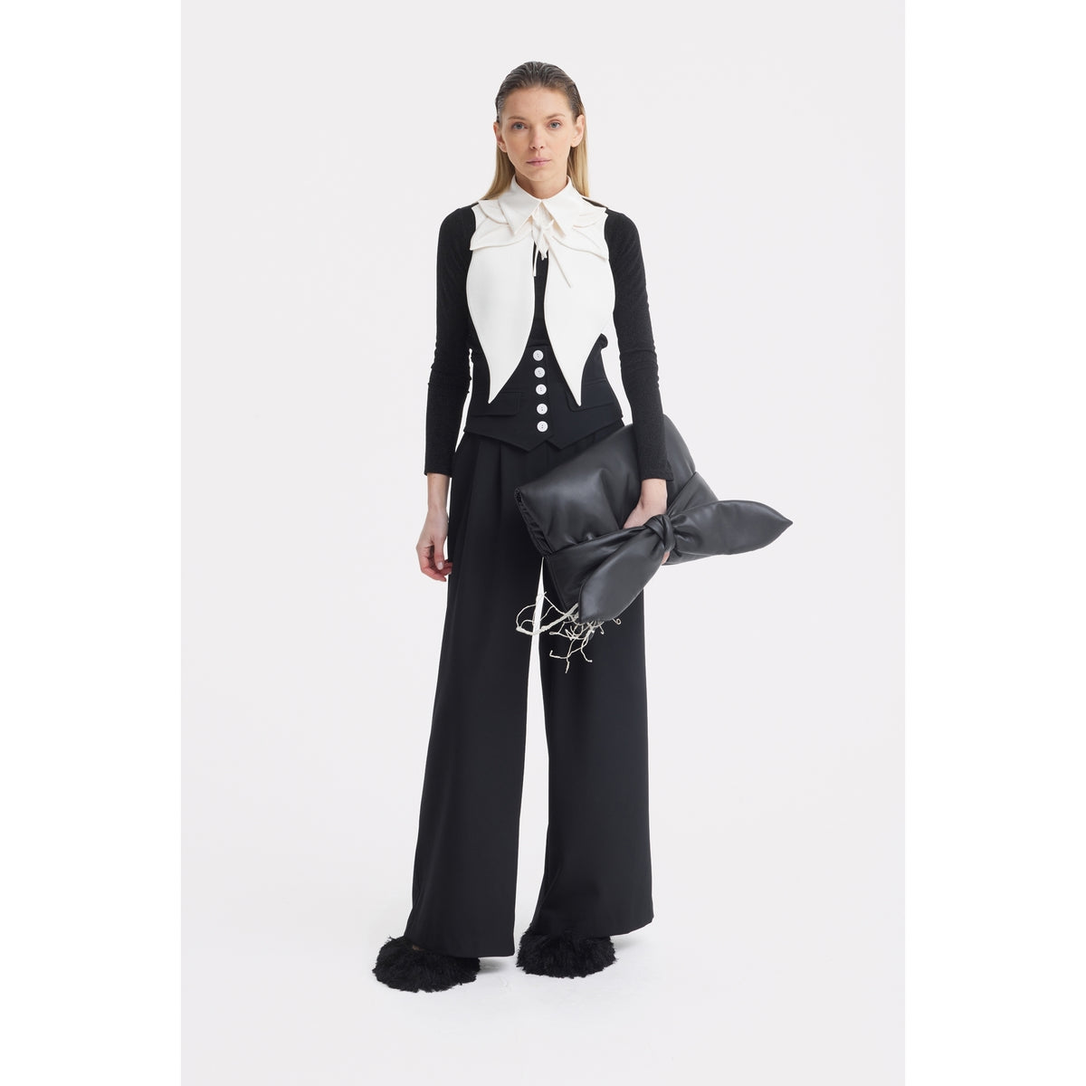 A woman with straight, slicked-back hair and a neutral expression wears JULIA ALLERT's Buttoned Corset-Waist Black Tailored Trousers, a black long-sleeve top, black and white vest, and holds a large black cushion-shaped bag against a white background.