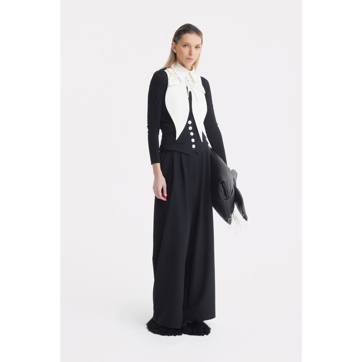 A woman stands against a plain white background wearing JULIA ALLERT's Buttoned Corset-Waist Black Tailored Trousers and a fitted top, holding a large black pillow-like accessory. Her straight hair is tucked behind her ears.