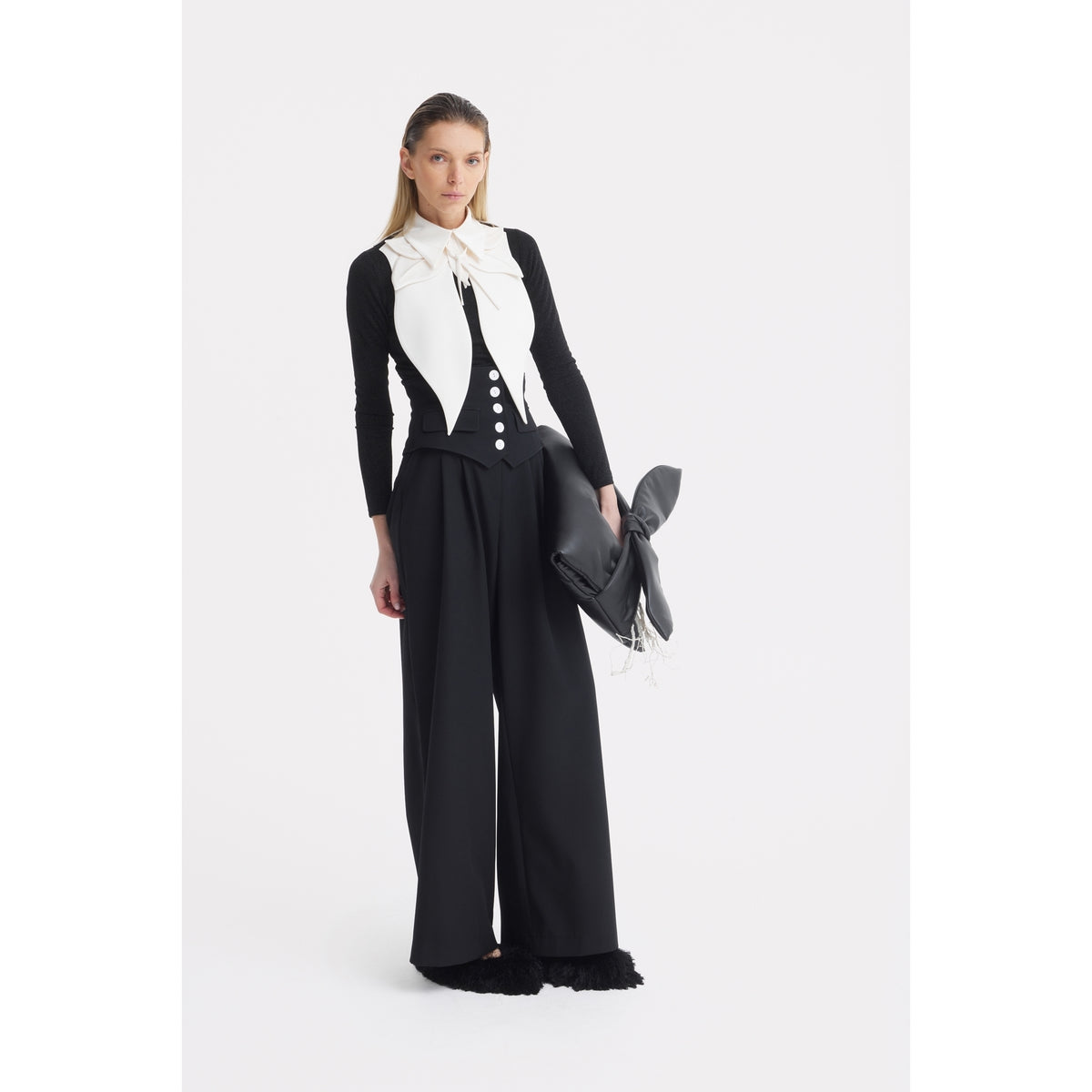 A woman stands against a white background in a black long-sleeve top, a fitted black vest with white buttons, and JULIA ALLERT Buttoned Corset-Waist Black Tailored Trousers, accessorizing with a large white bow scarf and a black bow-detailed bag.