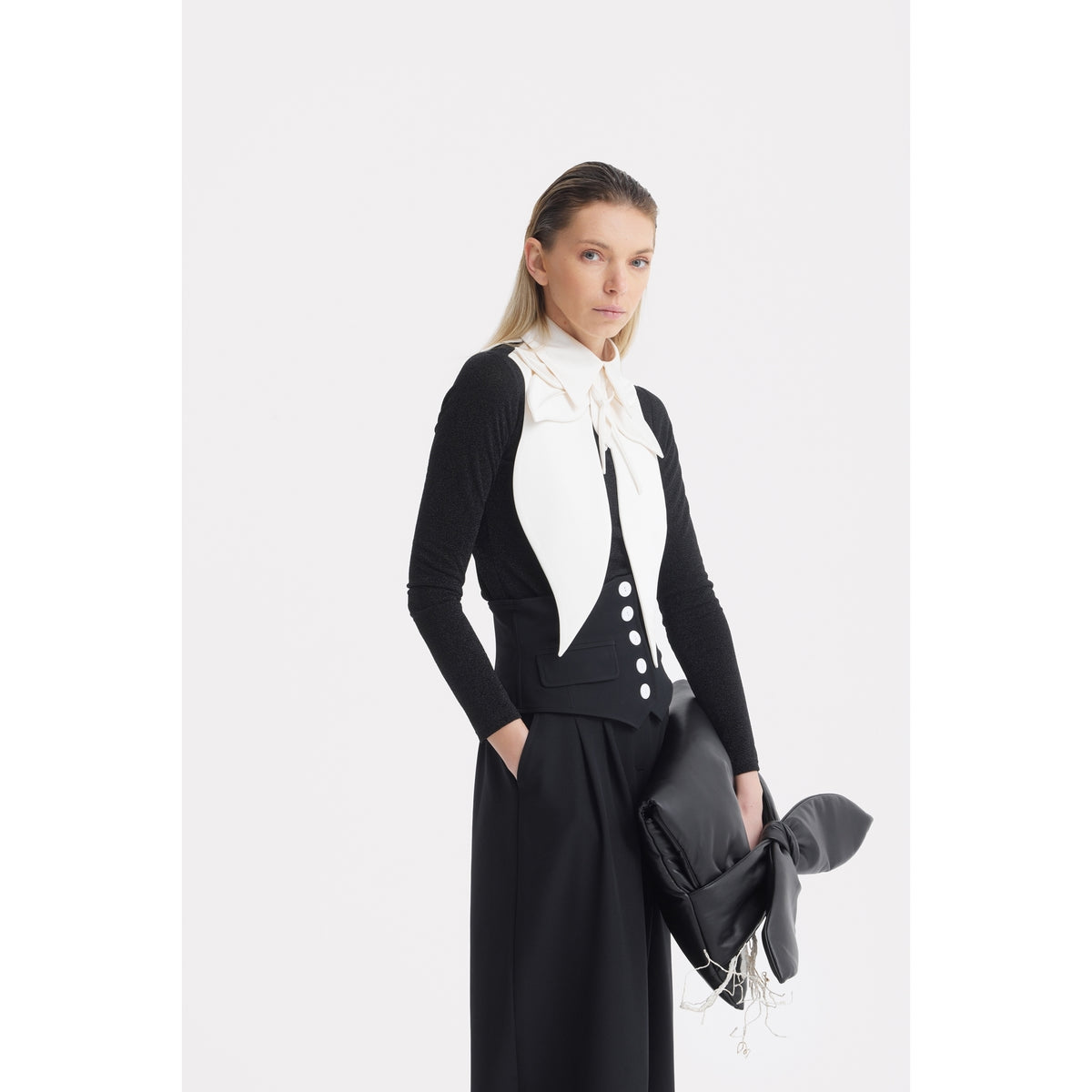 A woman in a black outfit with a large white bow collar holds a black bag with bow details, effortlessly pairing the look with JULIA ALLERT's Buttoned Corset-Waist Black Tailored Trousers.