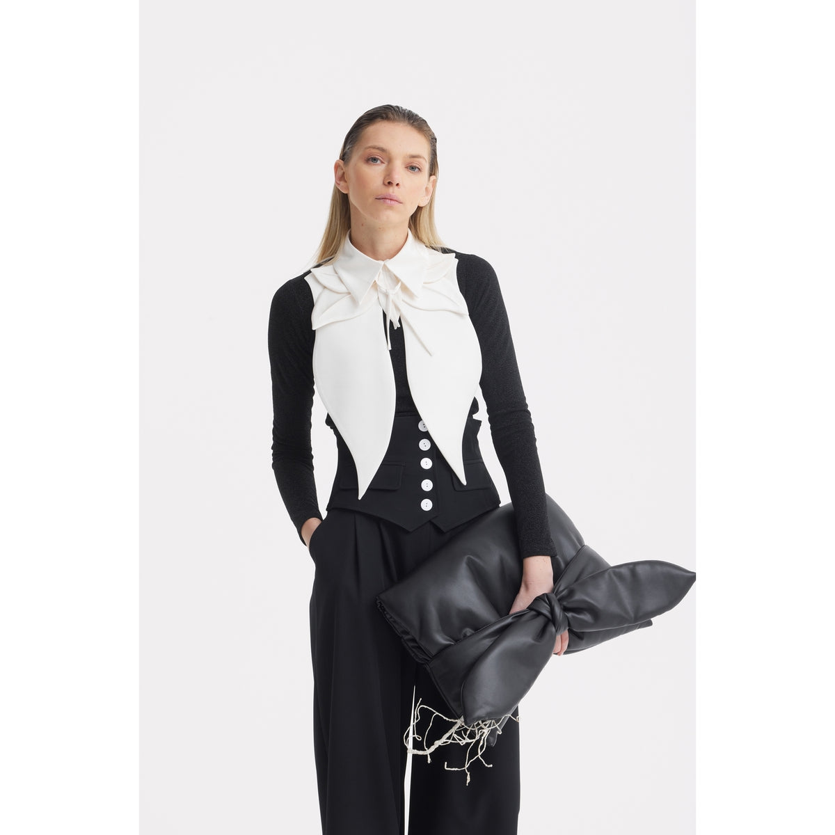 A woman stands against a white background in JULIA ALLERT Buttoned Corset-Waist Black Tailored Trousers, a black long-sleeve top, a white sleeveless vest with a large bow, and holds an oversized black clutch with string details.