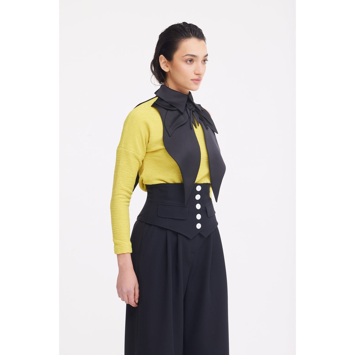 A woman with slicked-back dark hair stands against a white background, wearing a yellow long-sleeve top, high-waisted black pants with large front buttons, and the JULIA ALLERT Oversized Satin Bib Collar in black—a sculptural, bow-like accessory.