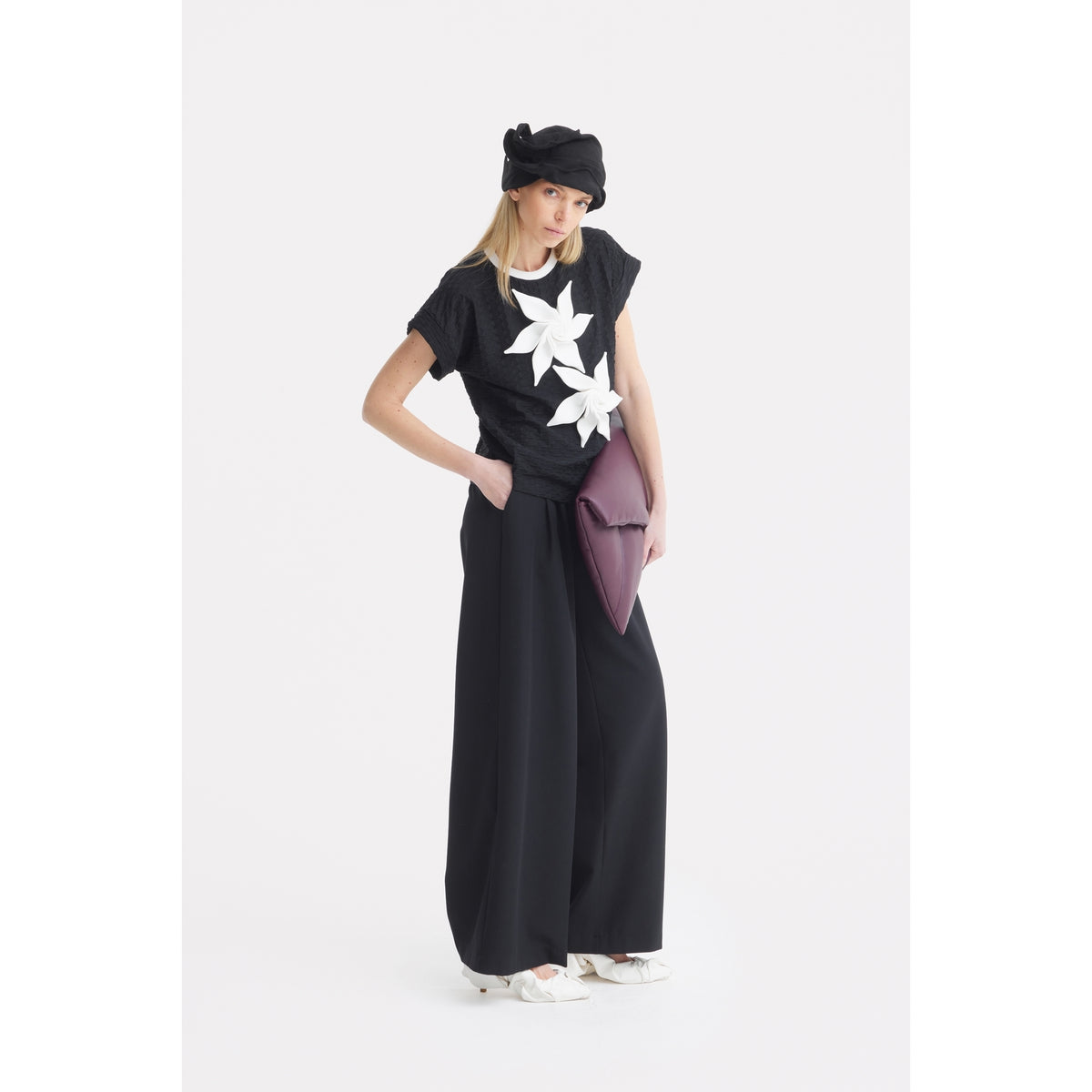 A woman in a JULIA ALLERT Black Short Sleeve Top With Floral Detail, wide-leg black pants, white sneakers, and holding a large purple clutch bag stands against a plain white background wearing a black beret.