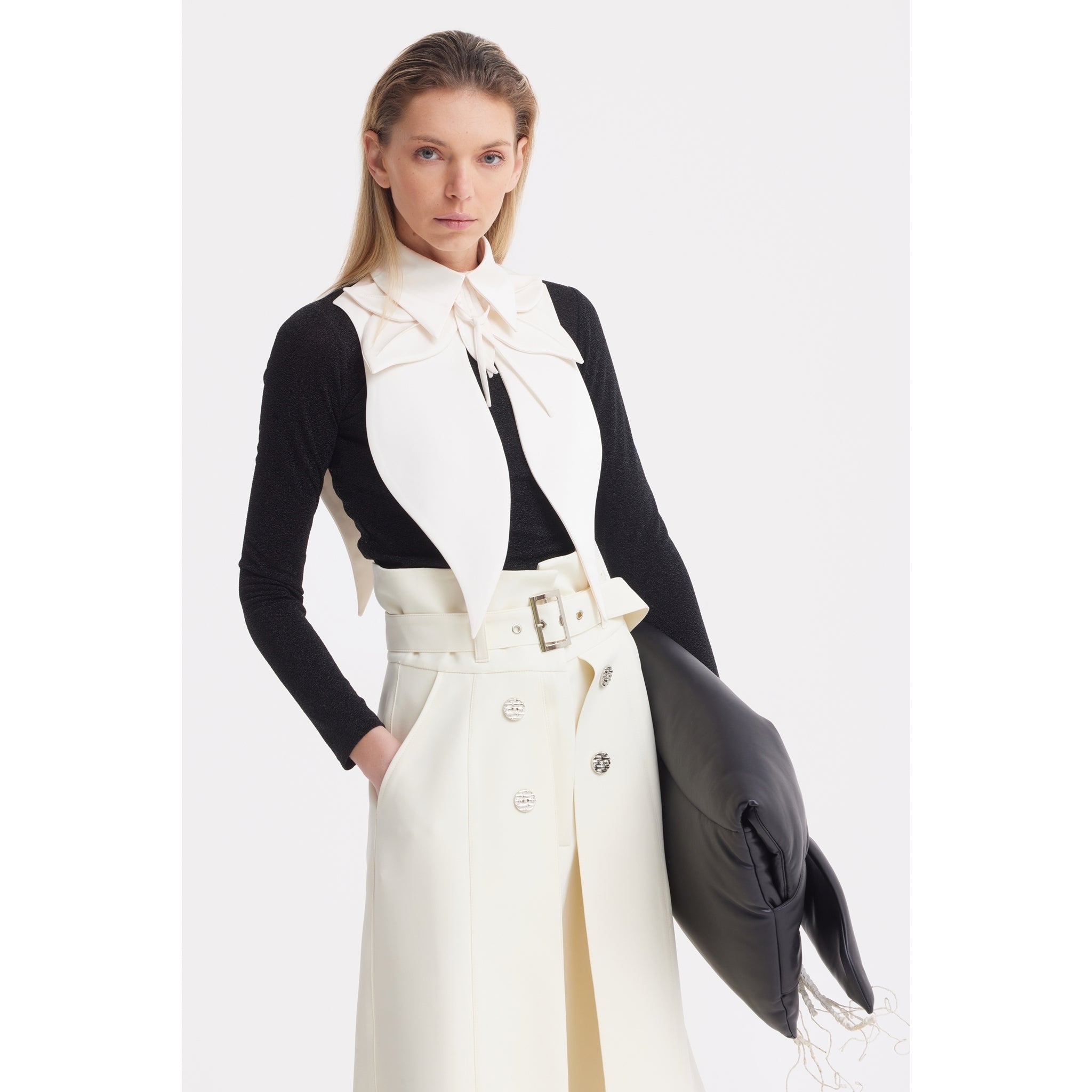 A woman wears a black long-sleeve top and white sleeveless vest featuring JULIA ALLERT's Oversized Satin Bib Collar in ivory, paired with a high-waisted white skirt and carrying a large black clutch under her arm.