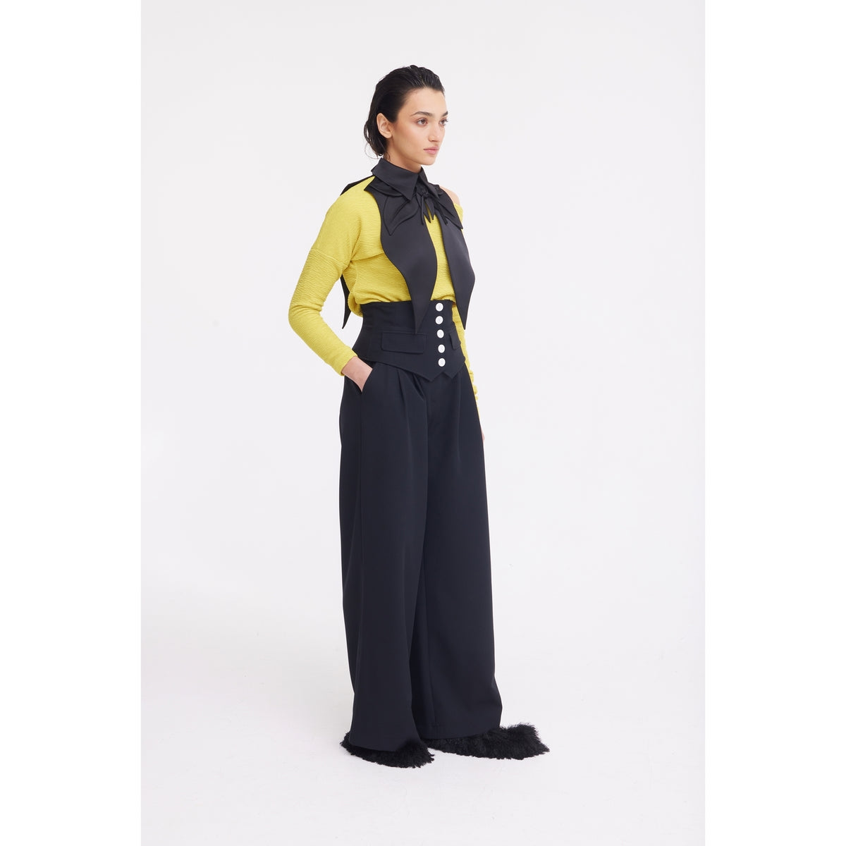A woman stands in a studio against a plain white background wearing JULIA ALLERT Buttoned Corset-Waist Black Tailored Trousers, a yellow long-sleeve top, a large black neck bow, and black furry shoes.
