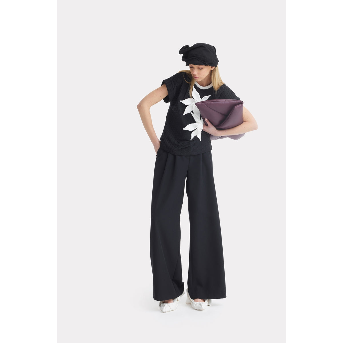 A woman in a black cap, wide-leg pants, and white shoes holds a large purple clutch while looking down. She's wearing the JULIA ALLERT Black Short Sleeve Top With Floral Detail. The background is plain white.