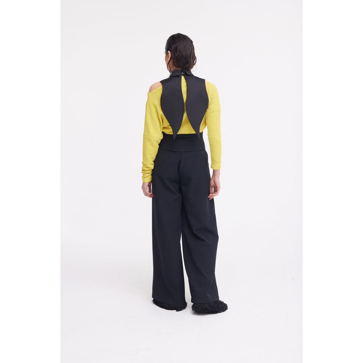 A person faces away, wearing a yellow cutout long-sleeve top, black vest with pointed back, JULIA ALLERT Oversized Satin Bib Collar - Black, and high-waisted wide-leg black pants against a plain white background.