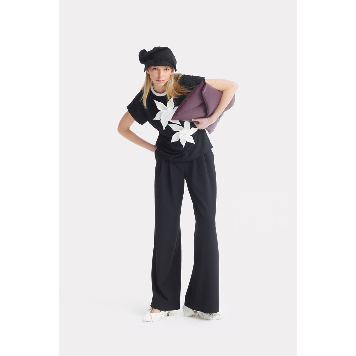 A woman in a JULIA ALLERT Black Short Sleeve Top With Floral Detail and wide-leg black pants poses, bending slightly forward. She accessorizes with a black hat, large purple bag, and white pointed shoes.