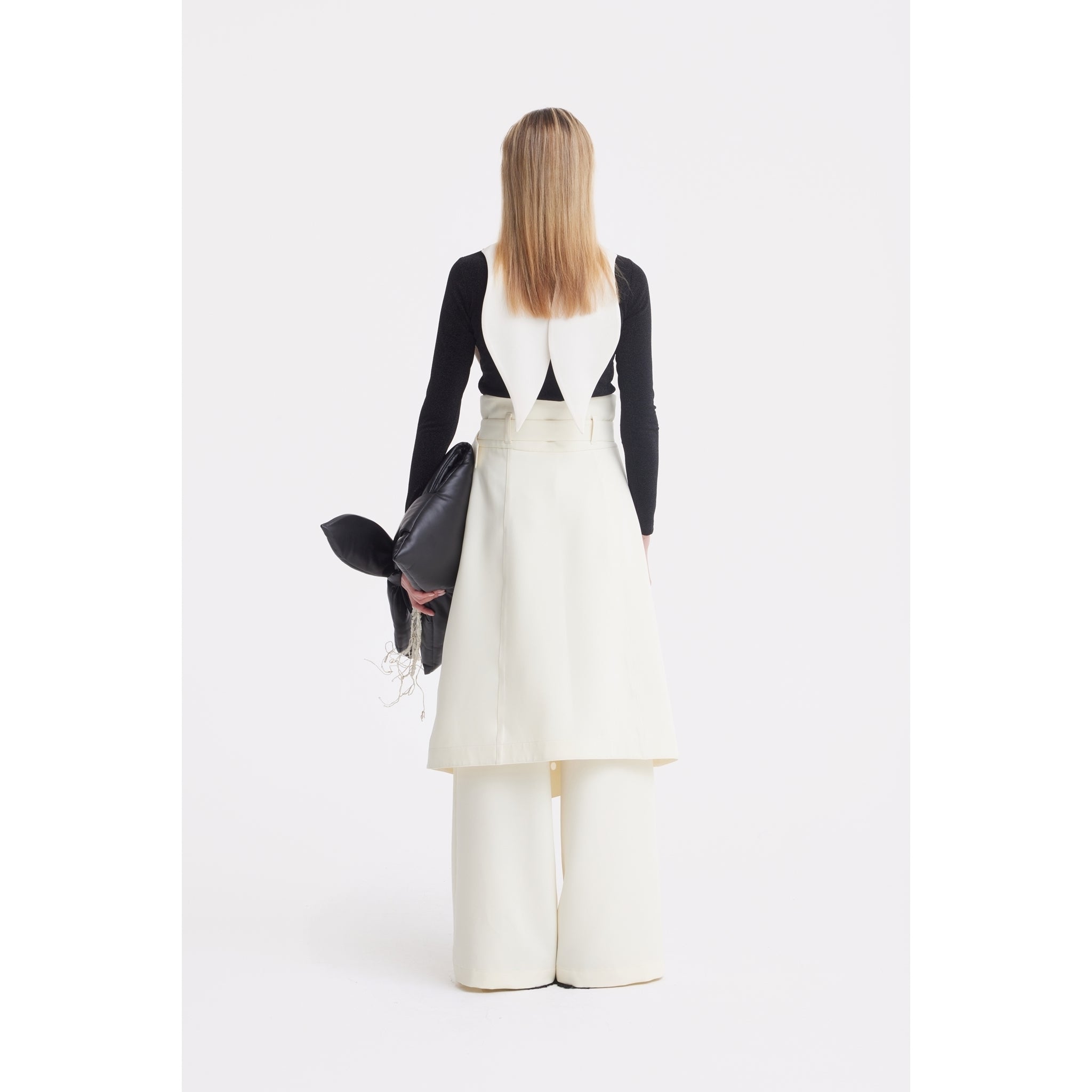 A person with straight, shoulder-length hair faces away, wearing the JULIA ALLERT Oversized Satin Bib Collar in Ivory over a black long-sleeve top and white sleeveless dress with wide-leg pants, holding a black bag against a white background.