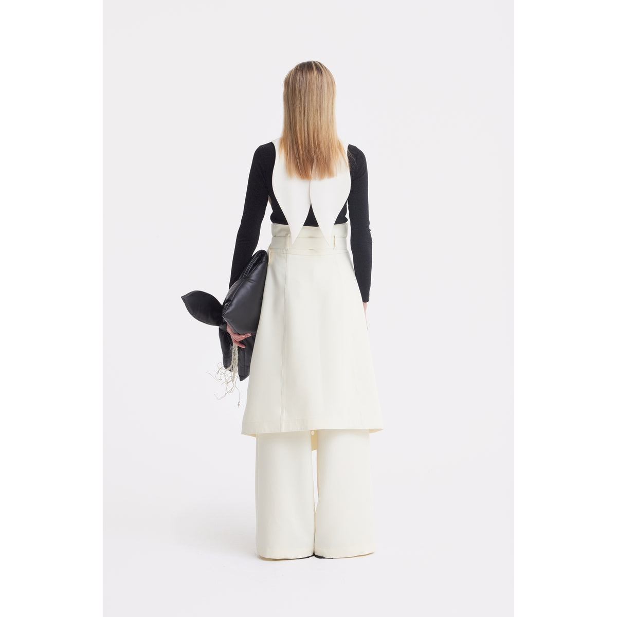 A person with straight, shoulder-length hair faces away, wearing the JULIA ALLERT Oversized Satin Bib Collar in Ivory over a black long-sleeve top and white sleeveless dress with wide-leg pants, holding a black bag against a white background.