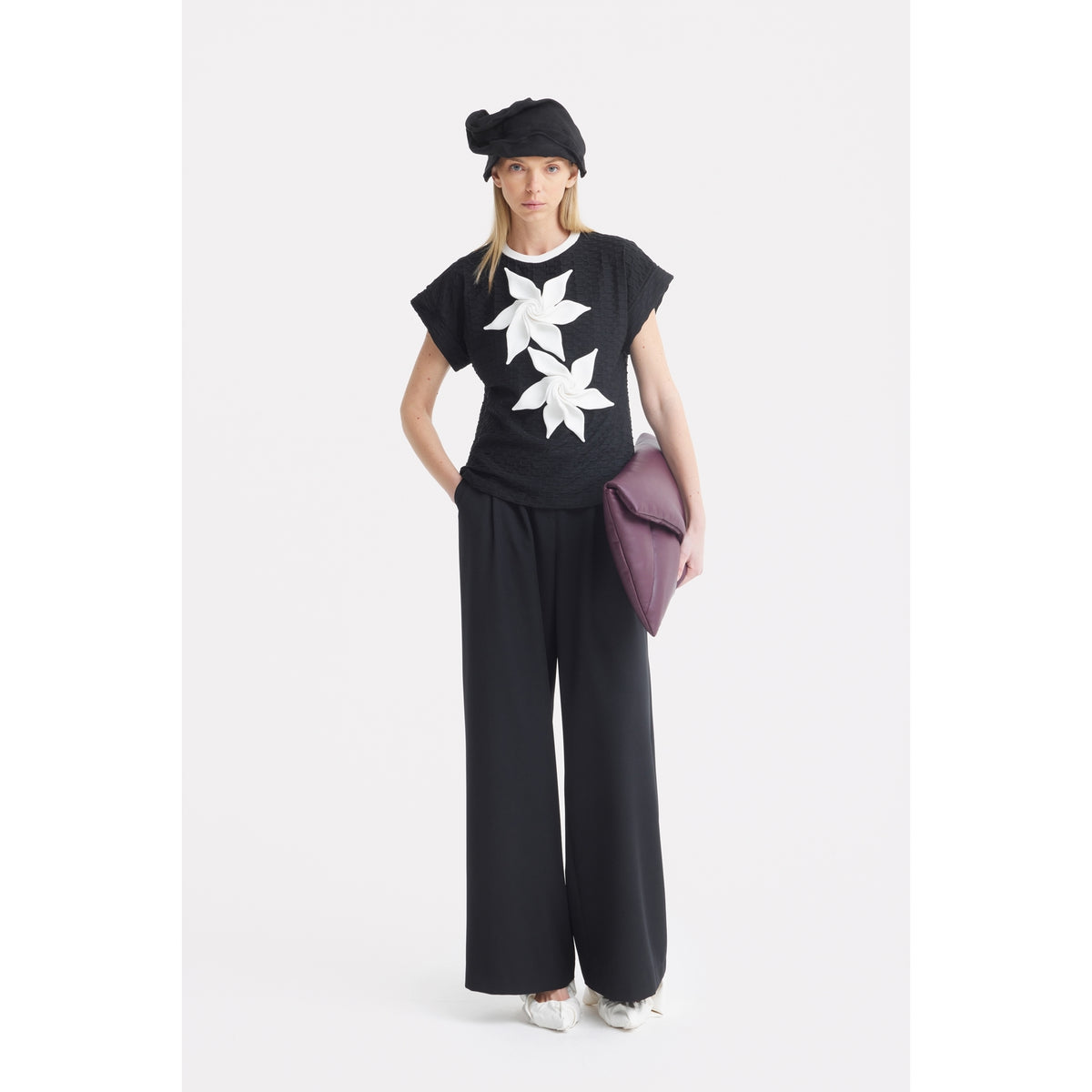 A woman faces forward in a JULIA ALLERT Black Short Sleeve Top With Floral Detail, wide-leg black pants, white shoes, a black hat, and holds a large purple clutch against a plain white background.