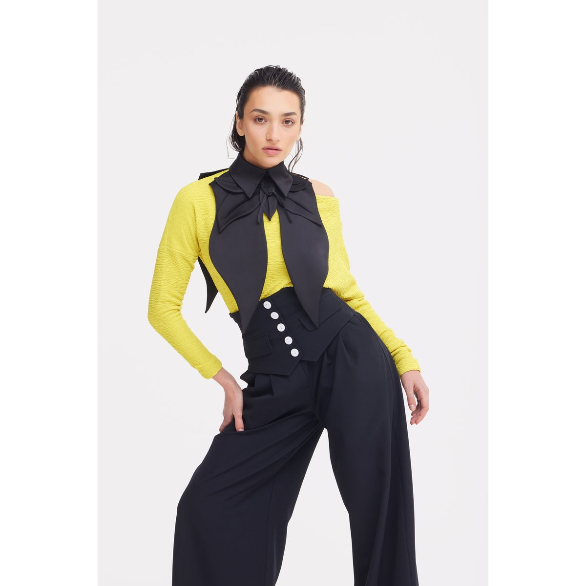 A woman with long dark hair poses confidently against a white background, wearing wide-leg black trousers, a yellow off-the-shoulder top, and the JULIA ALLERT Oversized Satin Bib Collar in black as a sculptural accessory.
