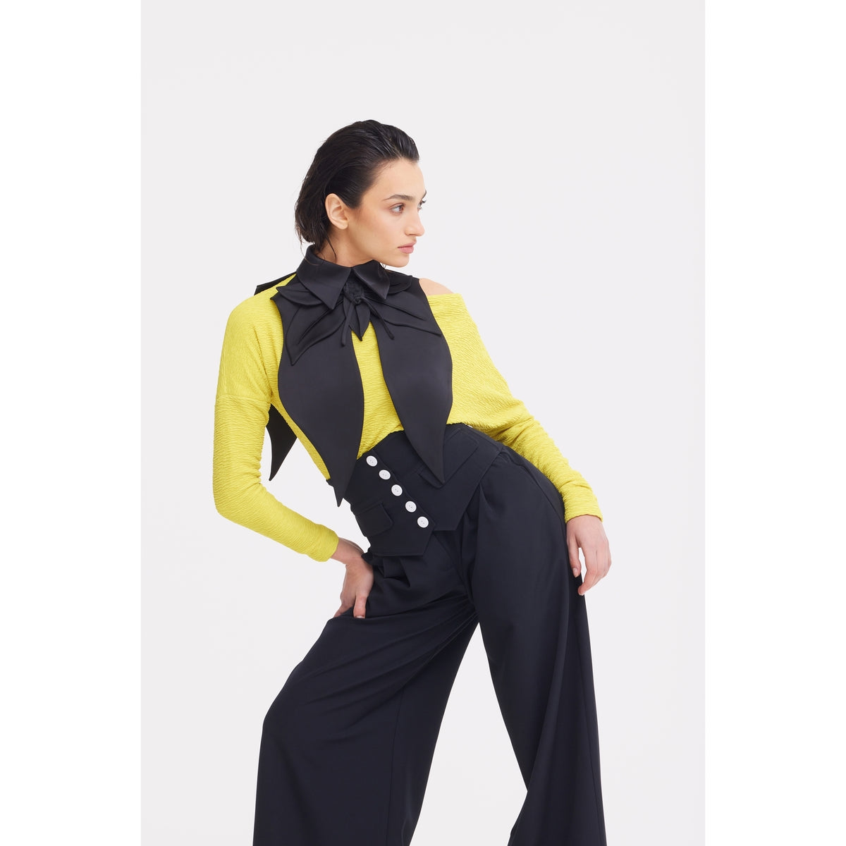 A woman poses against a plain white background wearing JULIA ALLERT Buttoned Corset-Waist Black Tailored Trousers, styled with a yellow top, black vest with white buttons, and a large black bow at the collar.