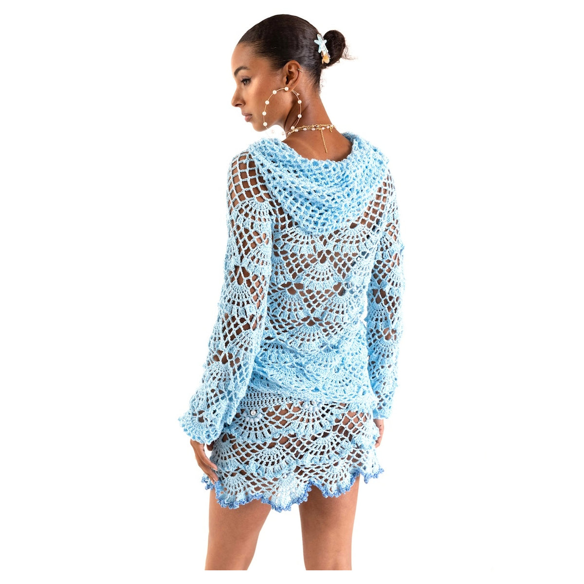 A woman in a light blue crochet top and the ANDREEVA Aqua Marine Handmade Mini Crochet Skirt with scalloped edges looks over her shoulder, accessorized with hoop earrings, a necklace, and a flower hair clip against a white background.