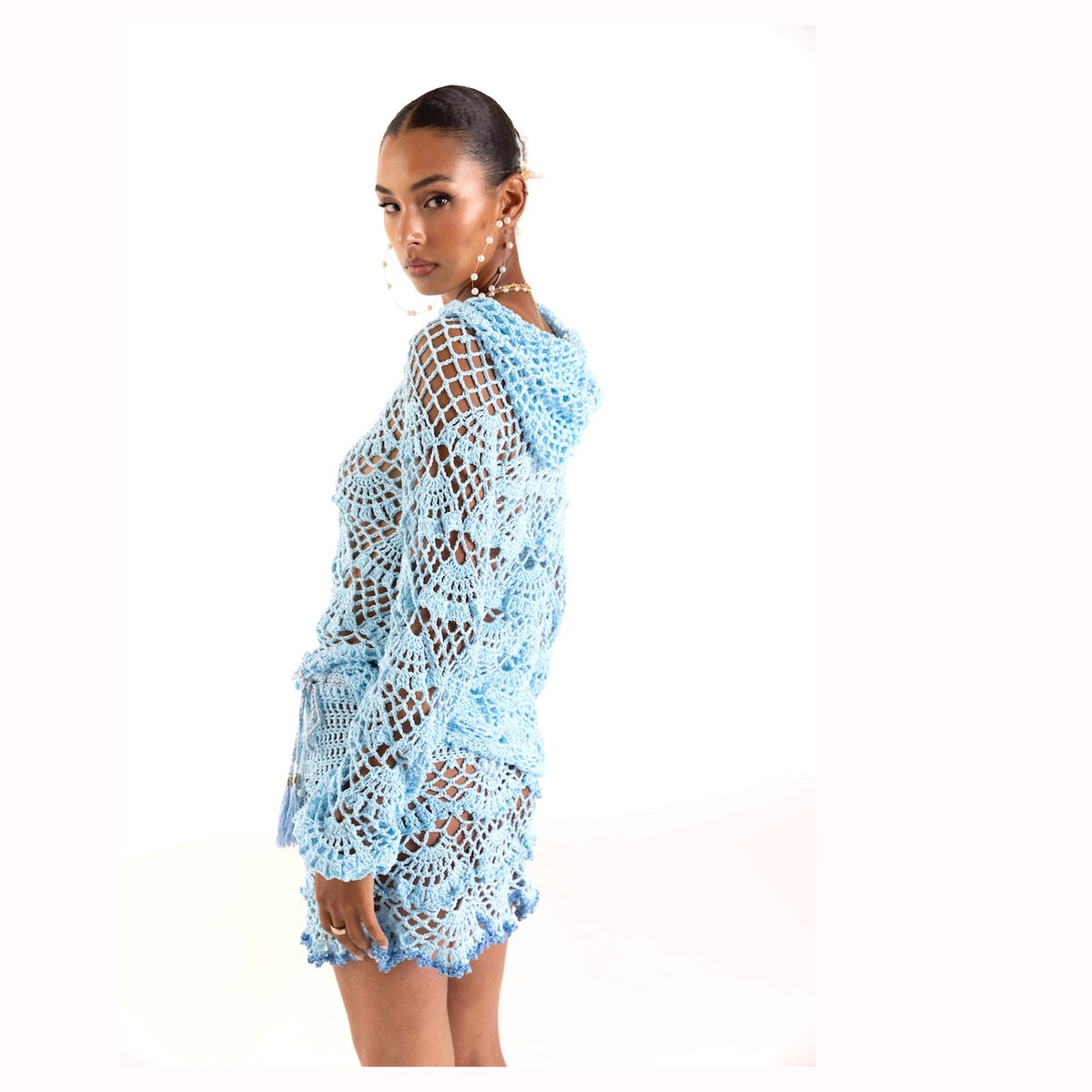 A woman poses sideways against a white background in the ANDREEVA Aqua Marine Handmade Crochet Hoodie, a light blue open-weave dress with long sleeves. She wears pearl earrings and has her hair pulled back.
