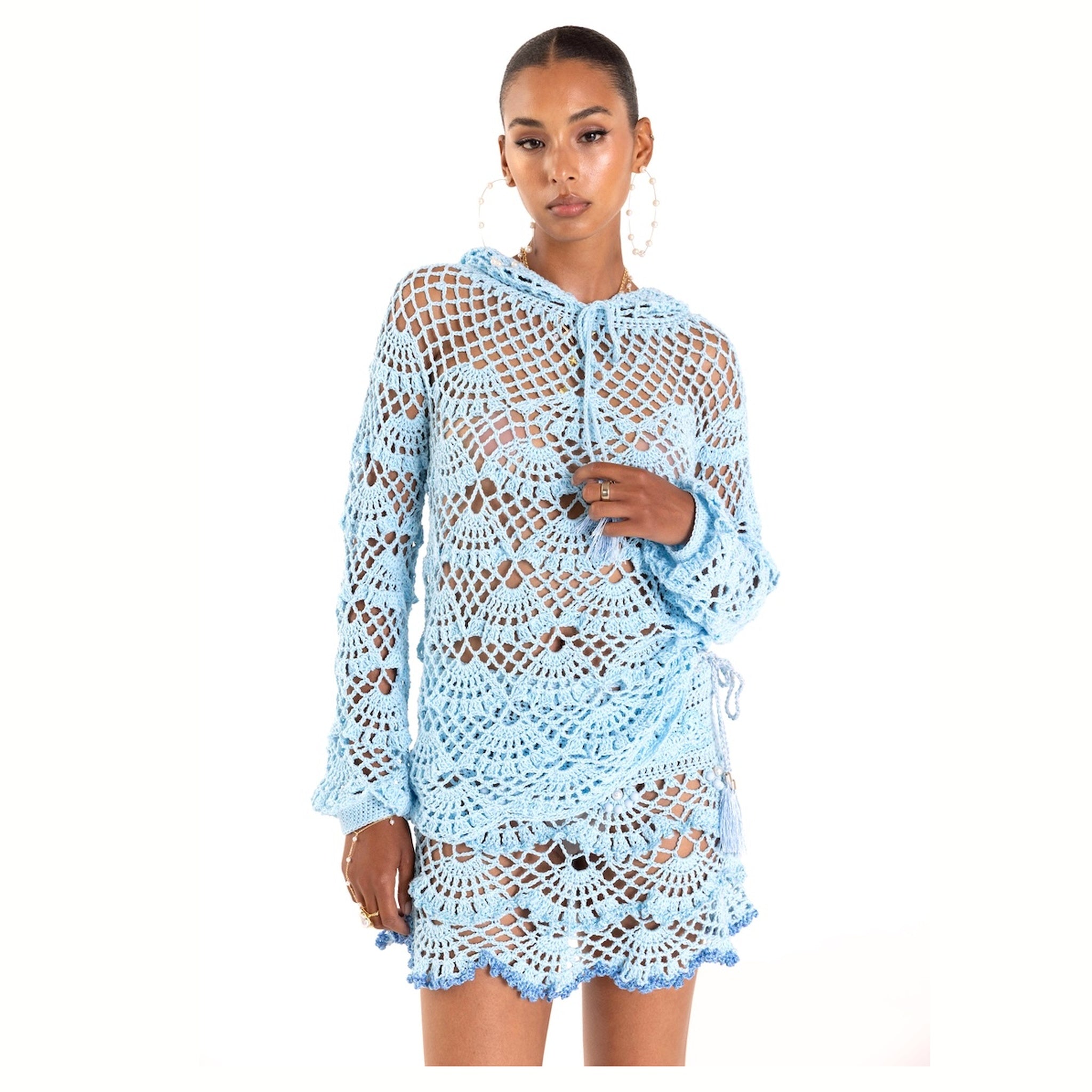 A woman models the ANDREEVA Aqua Marine Handmade Mini Crochet Skirt, a light blue, semi-sheer piece with an intricate open-knit pattern and scalloped edges—ideal for beachwear—as she poses against a white backdrop.