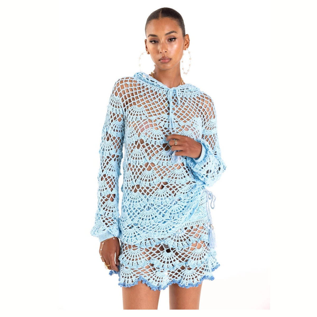 A woman models the ANDREEVA Aqua Marine Handmade Mini Crochet Skirt, a light blue, semi-sheer piece with an intricate open-knit pattern and scalloped edges—ideal for beachwear—as she poses against a white backdrop.