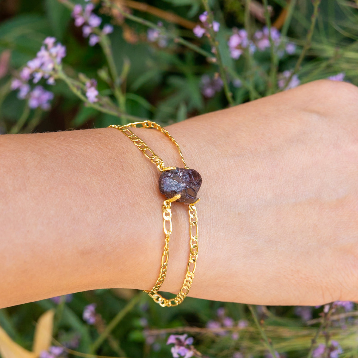 Gold chain bracelet with a dark stone on a wrist against a natural background with flowers.