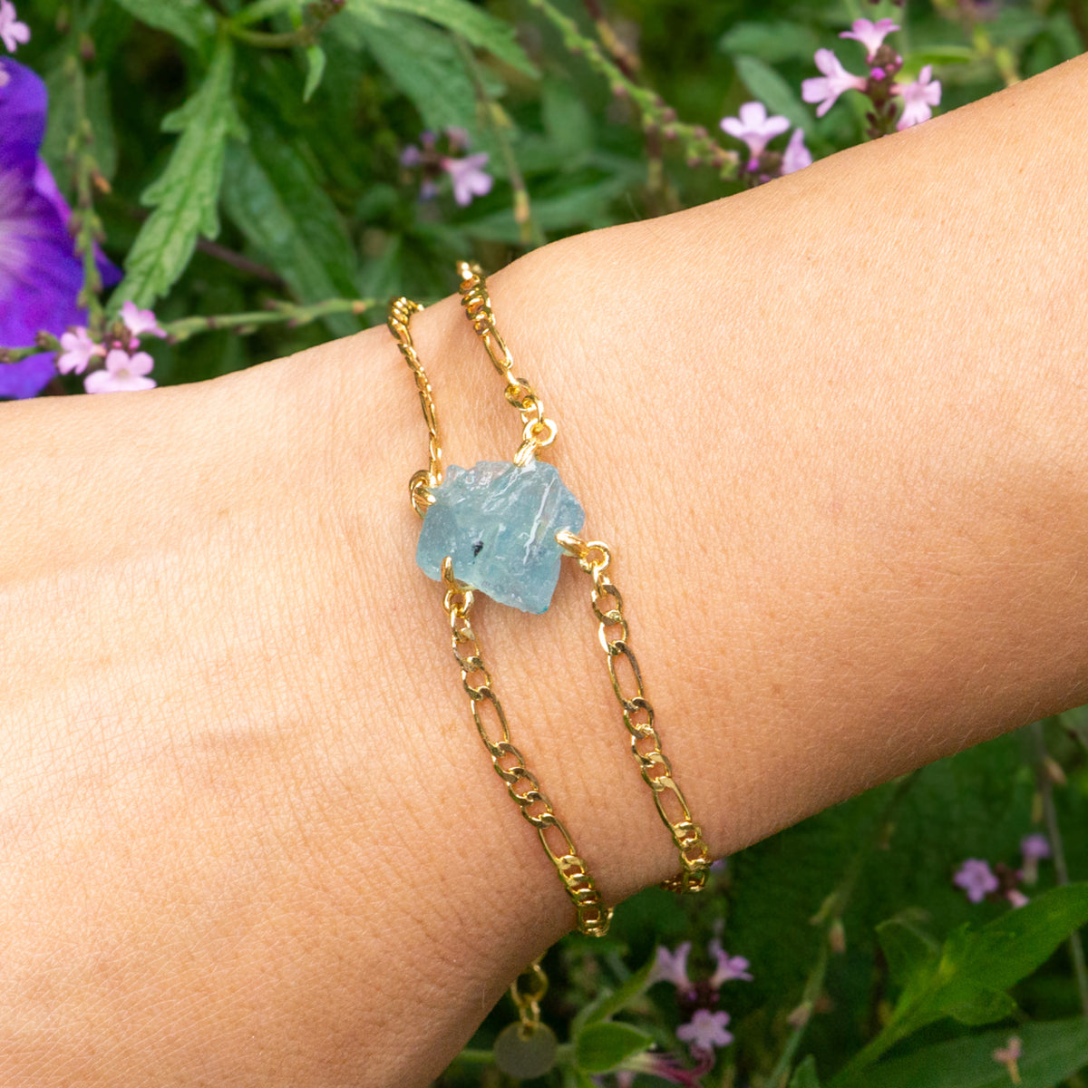 Gold bracelet with a blue stone on a wrist against a natural background with flowers.