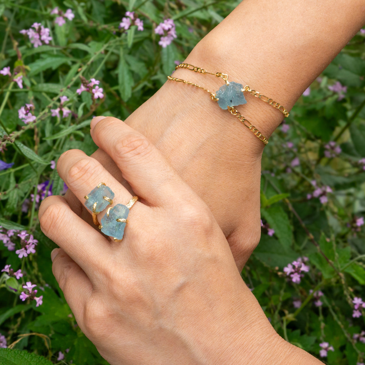 Hand wearing a gold bracelet and ring with blue stones against a green background with flowers
