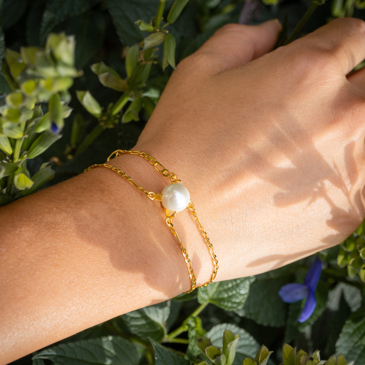 Gold bracelet with a pearl on a wrist against a green leafy background