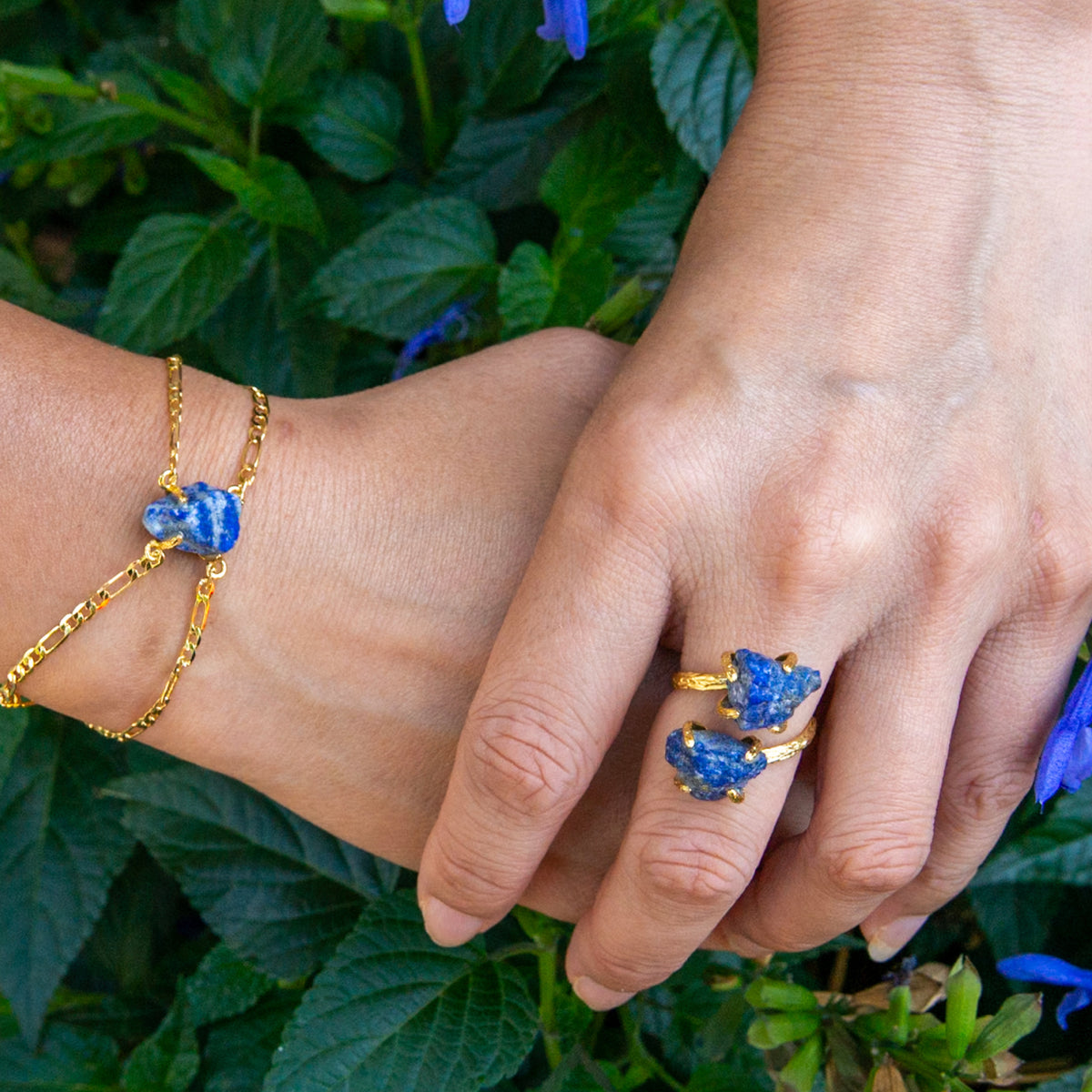 Close-up of hands wearing gold rings and bracelets with blue stones against a green leafy background
